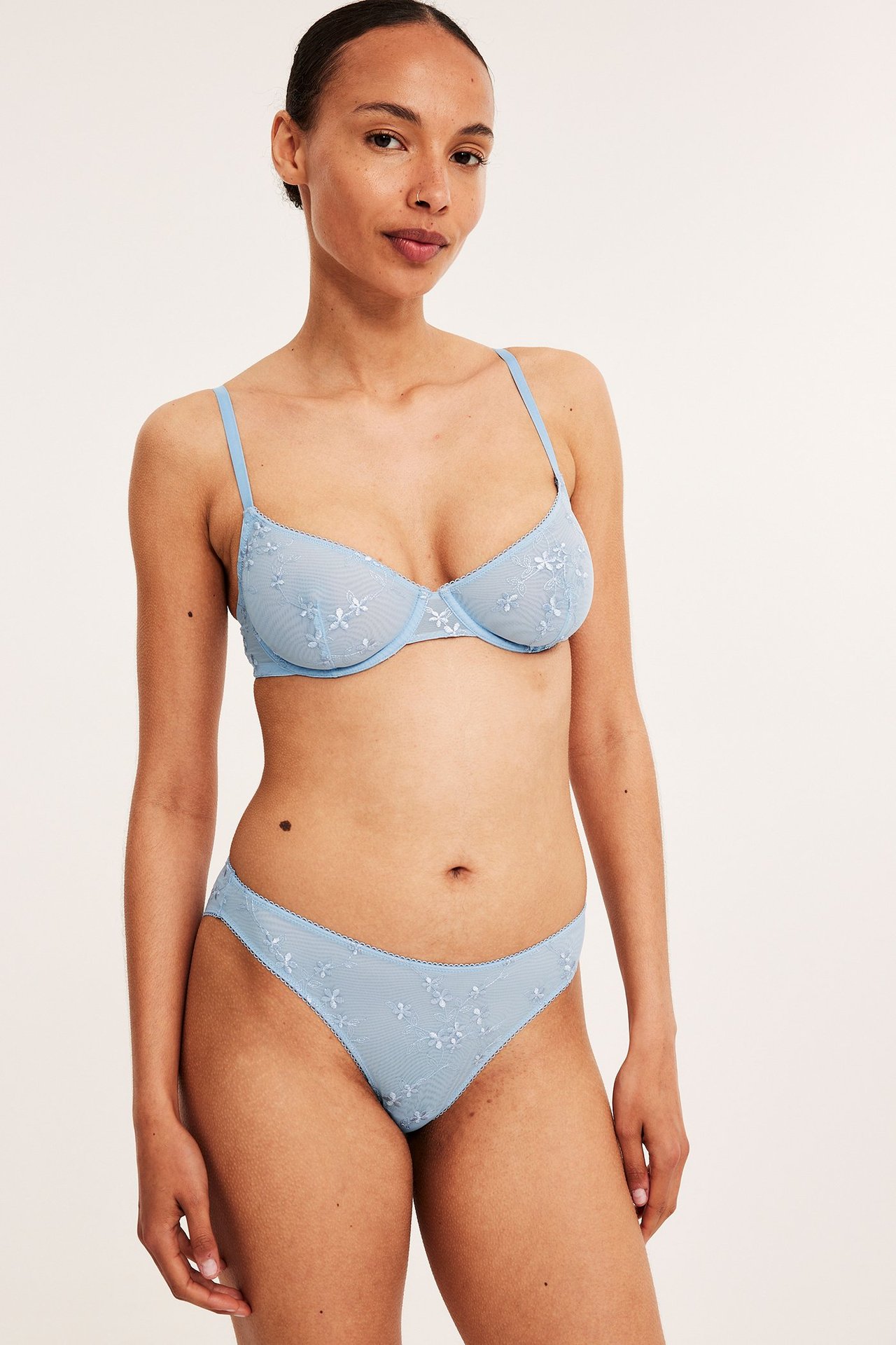 Framifrån, kvinna i ljusblå bygel-bh i mesh med blommig brodyr och matchande trosa.