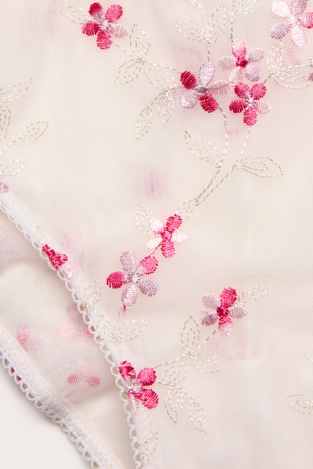 Närbild av offwhite skira brazilian trosor med rosa blommig brodyr och vågiga kanter.