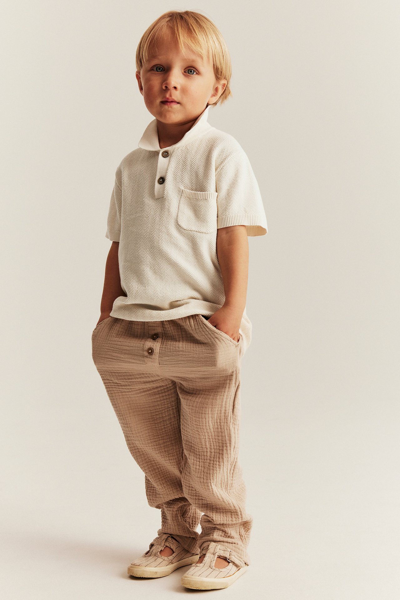 Widok z przodu dziecka w jasnobeżowej dzianinowej koszulce polo, jasnobrązowych spodniach i beżowych butach.