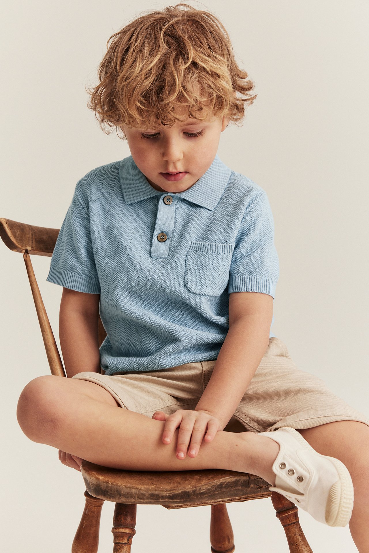 Forfra, et barn i lyseblå strikket pikétrøye med knapper og lomme, beige shorts og hvite tøysko.