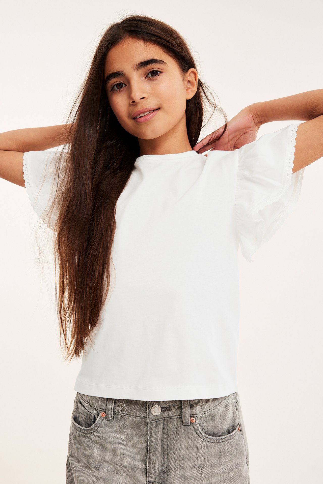 Frontvy av ett barn i en offwhite bomullstopp med volangärmar och grå jeans.