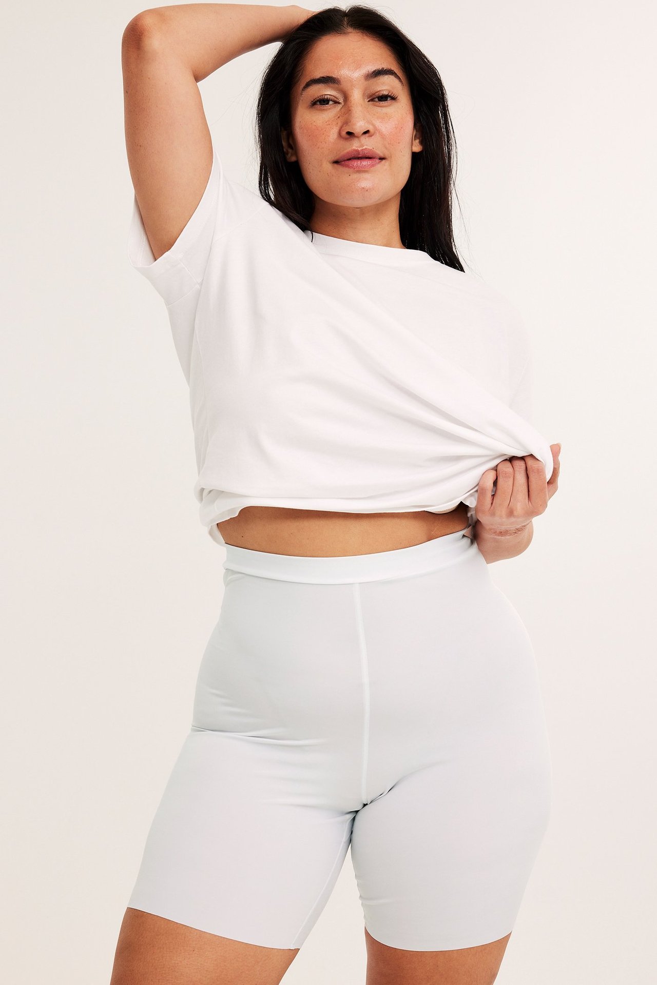 Forfra: kvinne i hvite, myke, tøyelige shaping-shorts og en hvit t-skjorte, trekker opp skjorten.