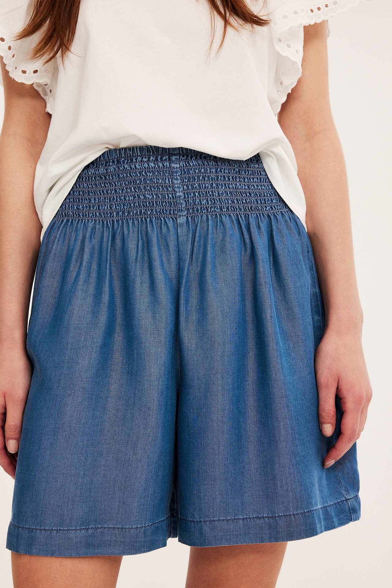 Nærbilde forfra av en kvinne i blå Tencel-shorts med smock i midjen og en hvit topp.