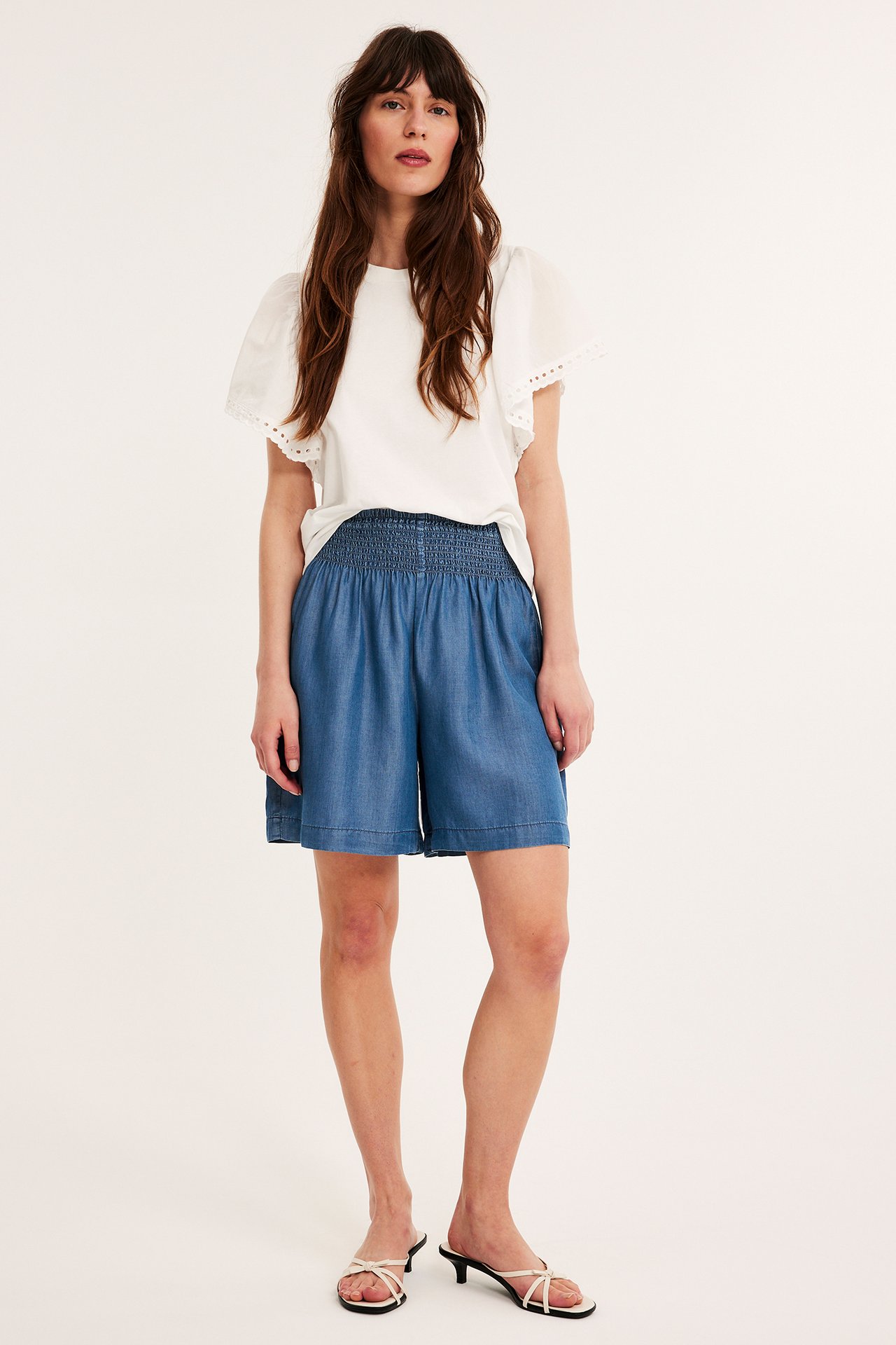 Frontbilde av kvinne i blå Tencel-shorts med smock i midjen, en hvit topp og hvite sandaler.