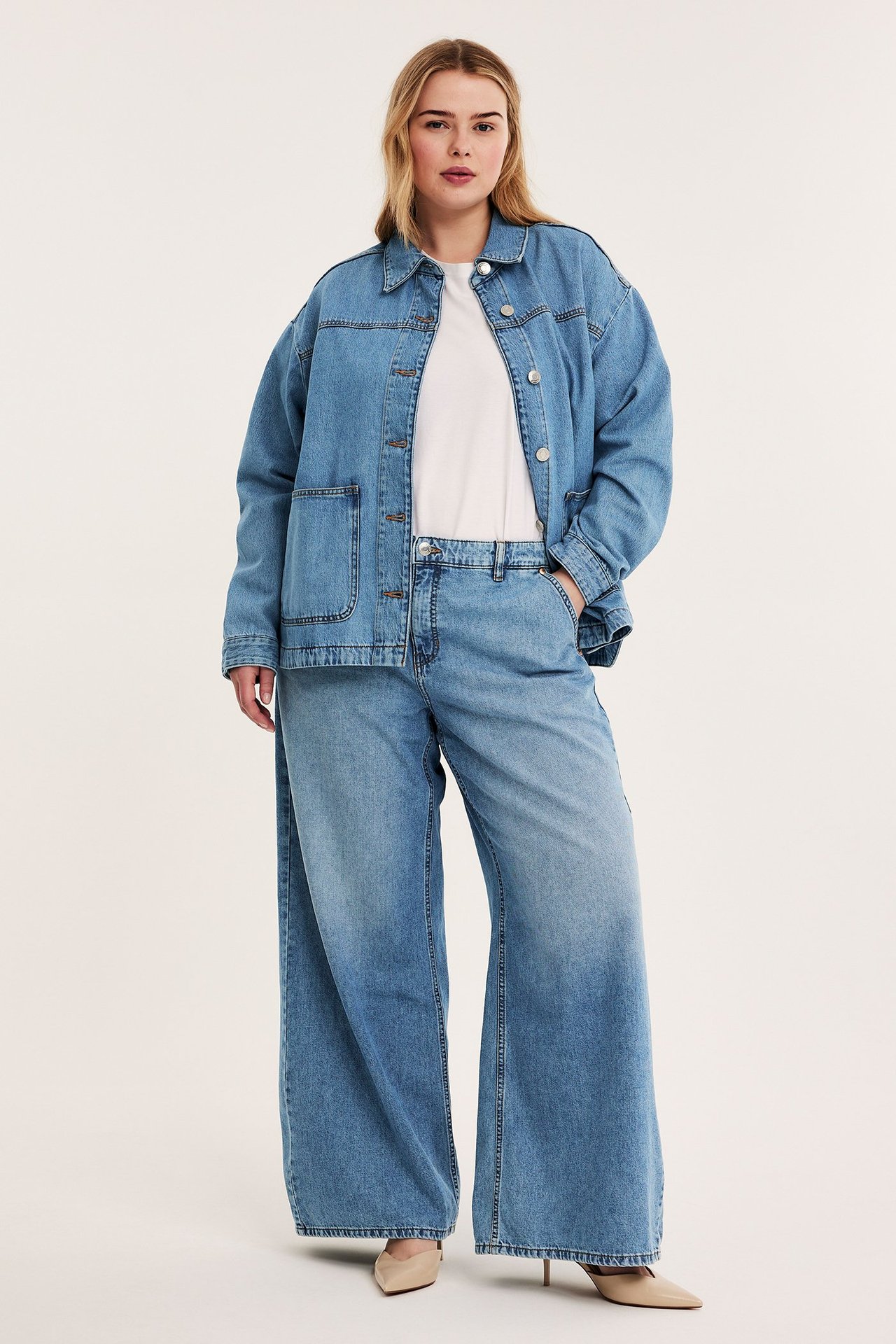 Framifrån: Kvinna i ljus denim vida jeans, matchande jeansjacka, vit t-shirt och beiga klackar.