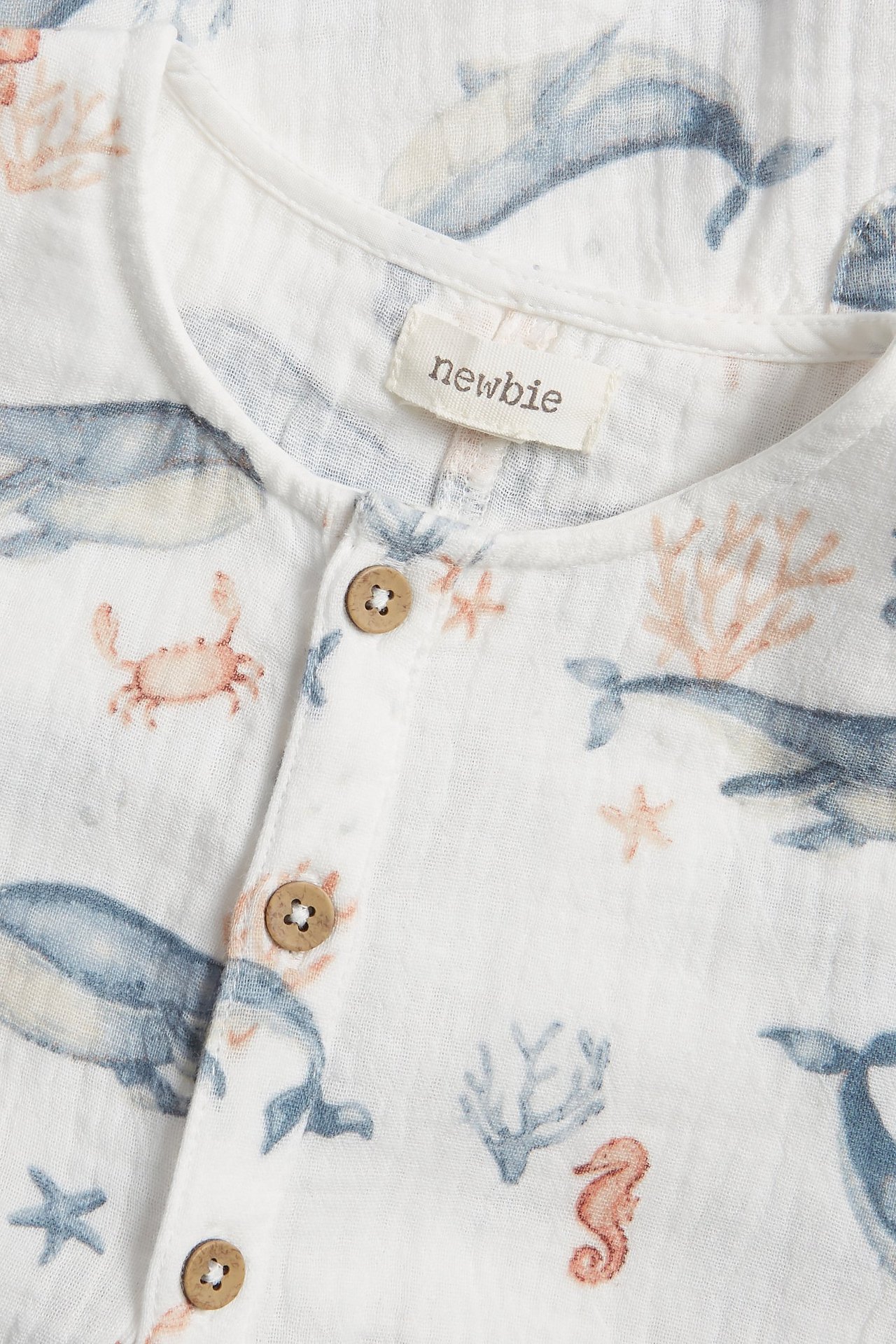 Nærbilde av en offwhite babyplaysuit i bomullsmuslin med blått sjødyrmønster og knapper foran.