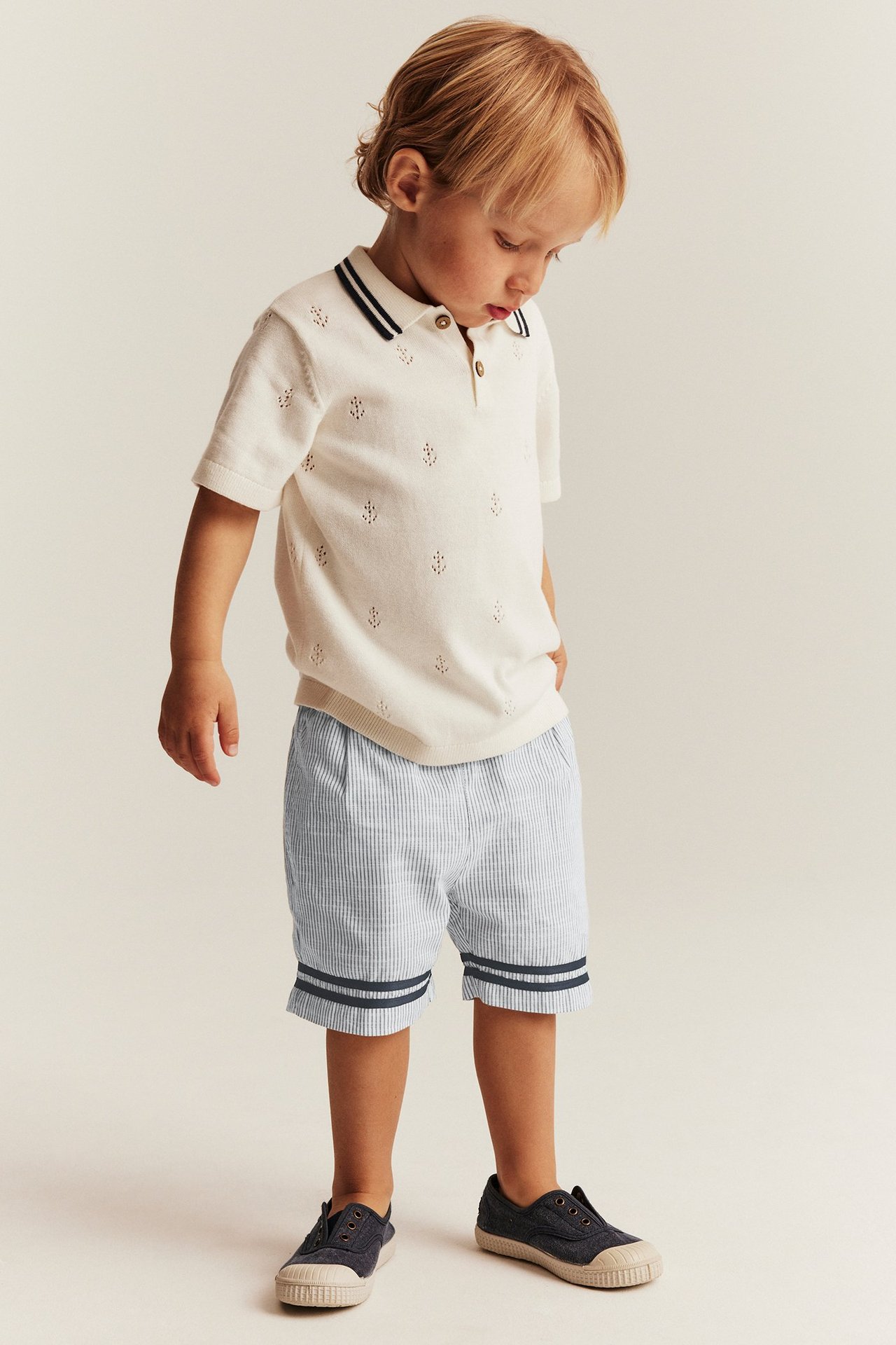 Kid i ljusblårandiga shorts med knappar, offwhite pikétröja och mörkblå canvasskor. Framifrån.