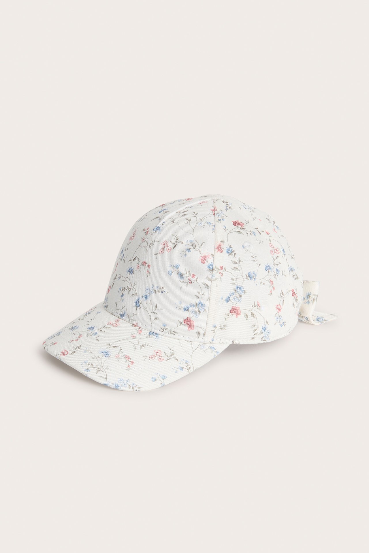 Sidevisning: Offwhite baby- eller barnekaps med blomstermønster og dekorativ sløyfe bak.