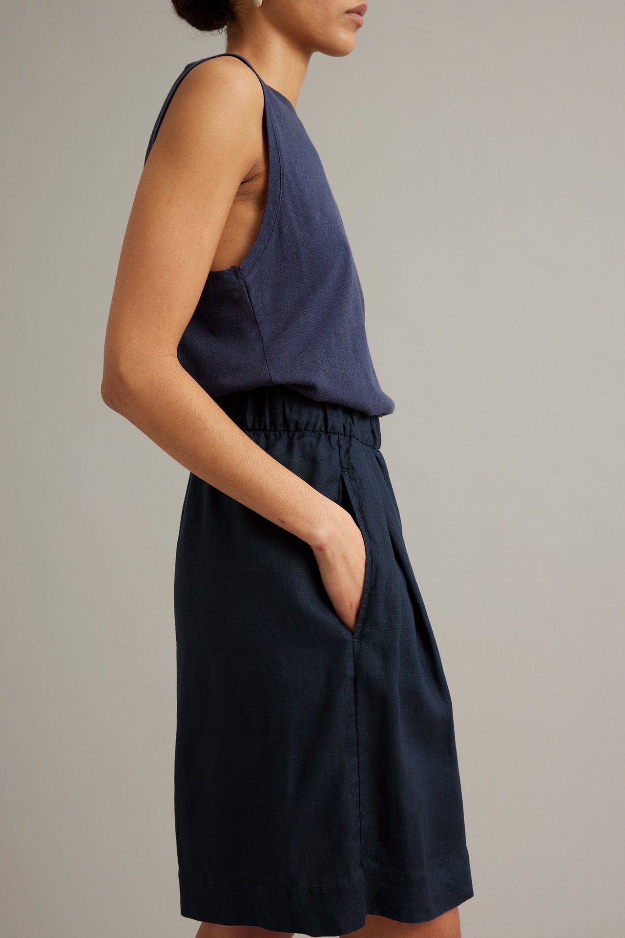 Nainen pukeutuneena tummansinisiin pellavaisiin polvipituisiin shortseihin, resori vyötäröllä, käsi taskussa ja yhteensopiva toppi.