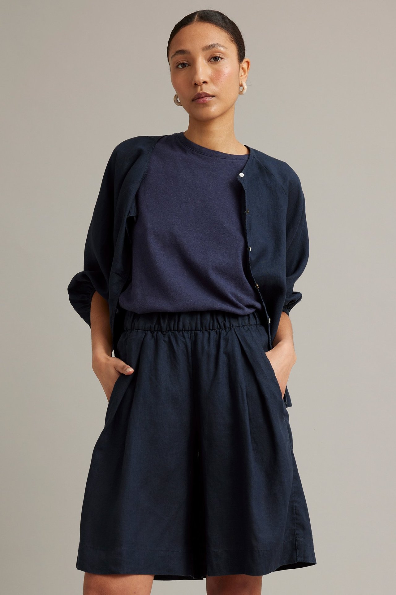 Edestä: Nainen tummansinisissä pellava-bermudashortseissa, tummansinisessä t-paidassa ja avonaisessa pellavatakkissa.