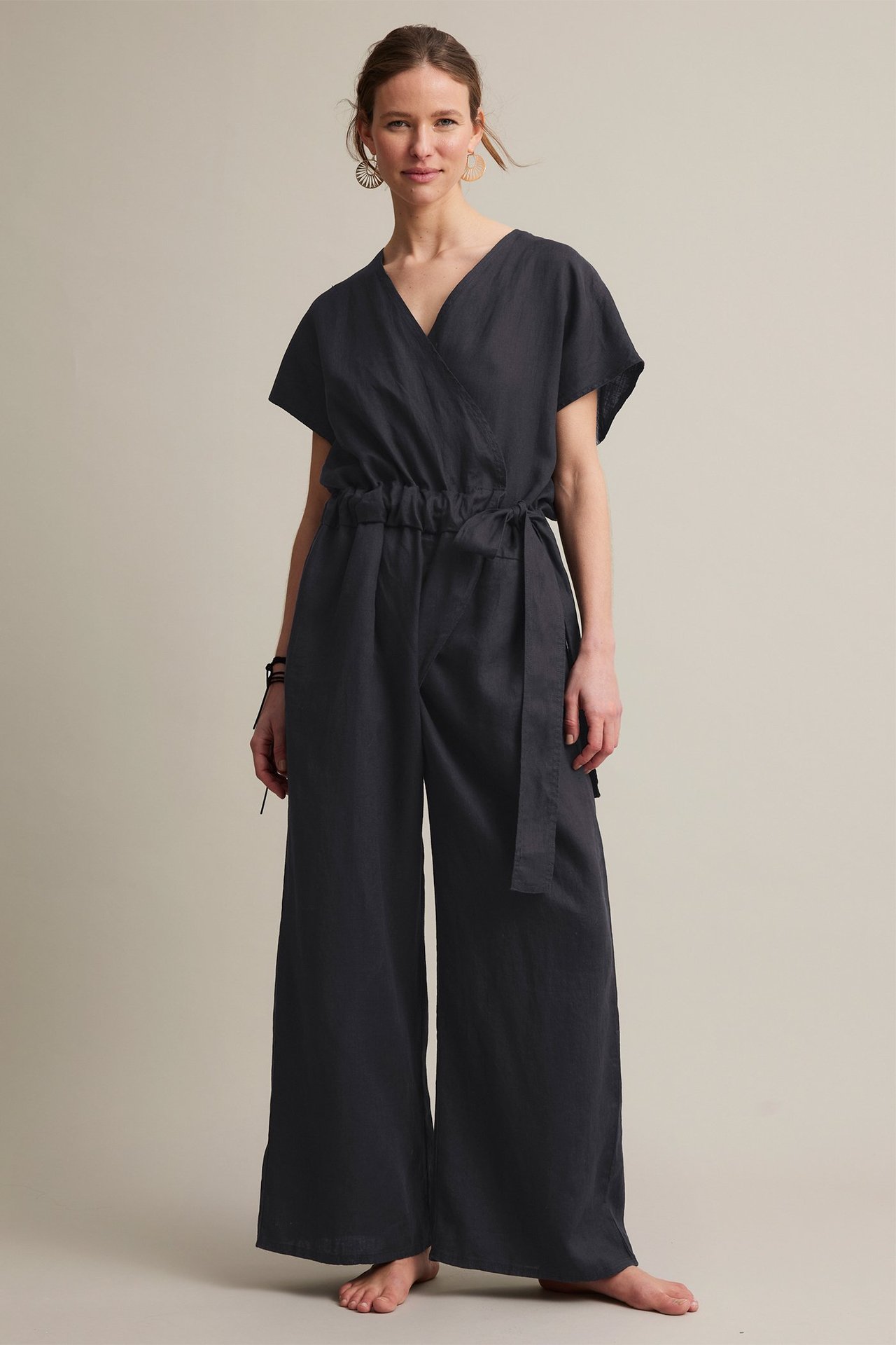 Nainen mustassa pellavahaalarissa, jossa kietaisuyksityiskohta, joustava vyötärö, leveät lahkeet ja viuhkakorvakorut.