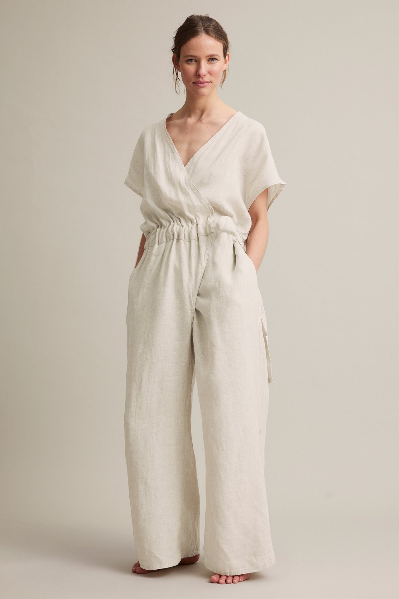 Forfra: kvinne i beige linnejumpsuit med omslagstopp, elastisk midje, vide ben, sidelommer.