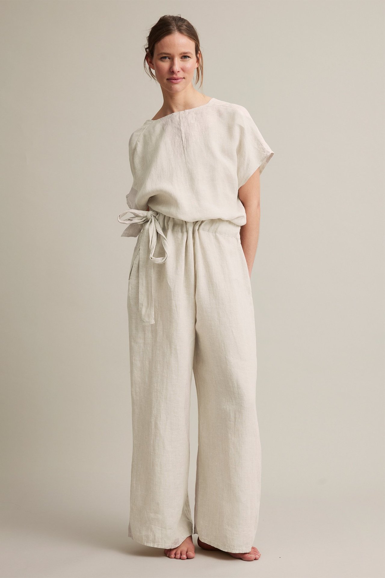Frontvisning: kvinne i beige linjumpsuit med omslagstopp, elastisk midje, knyting og rette ben.