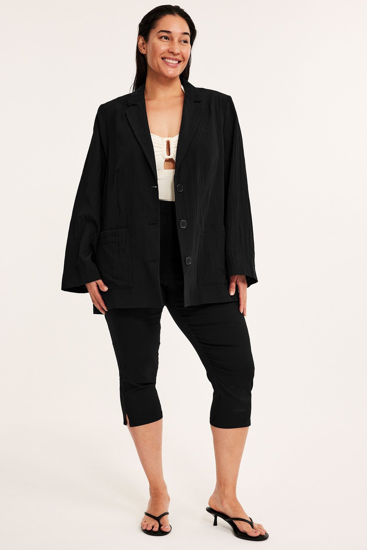 Kobieta w czarnych capri z rozcięciami, czarnej marynarce, kremowym topie i czarnych sandałach na obcasie, widok z przodu.