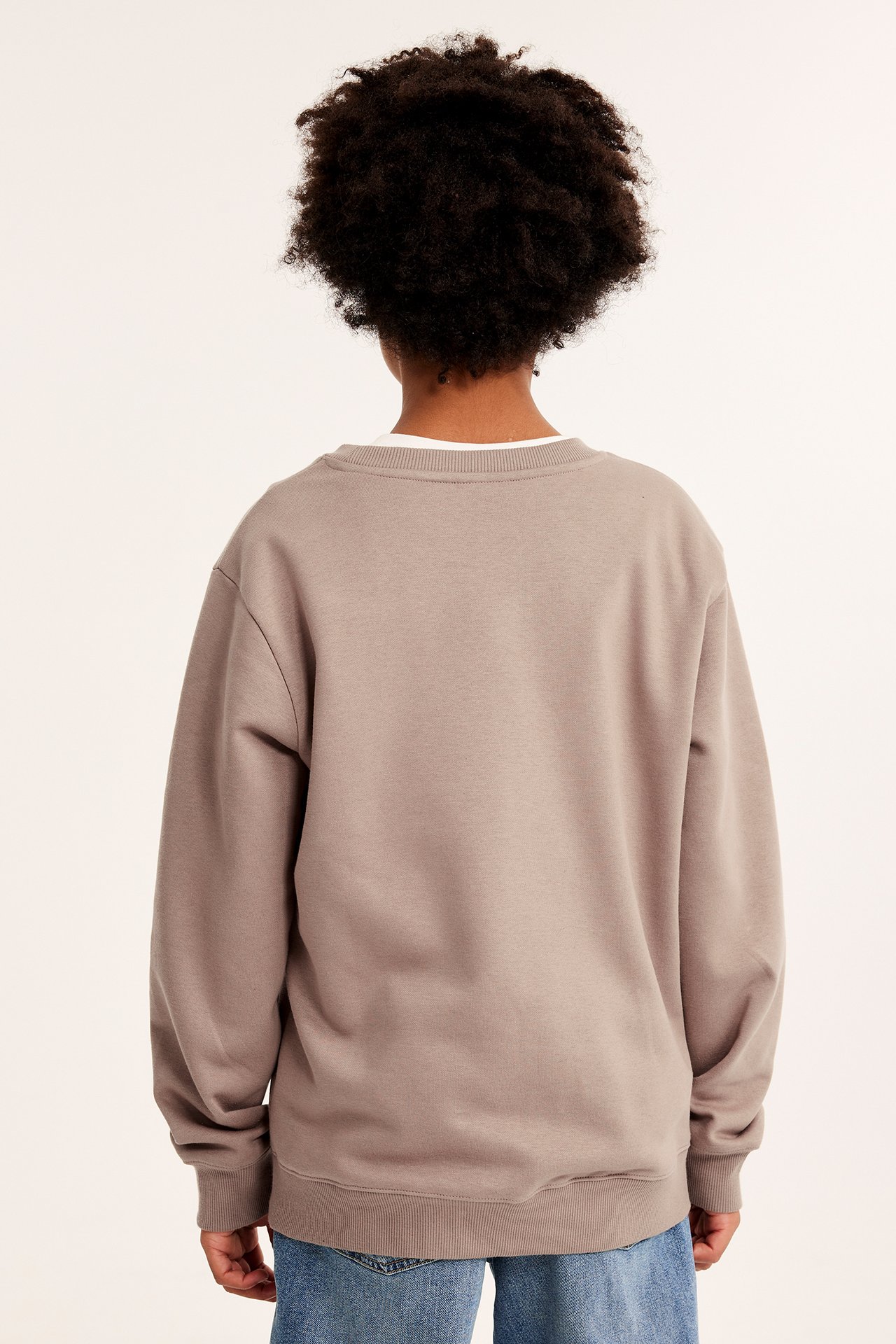 Bakfra av et barn i en brun, løs sweatshirt over en hvit t-skjorte og blå jeans.