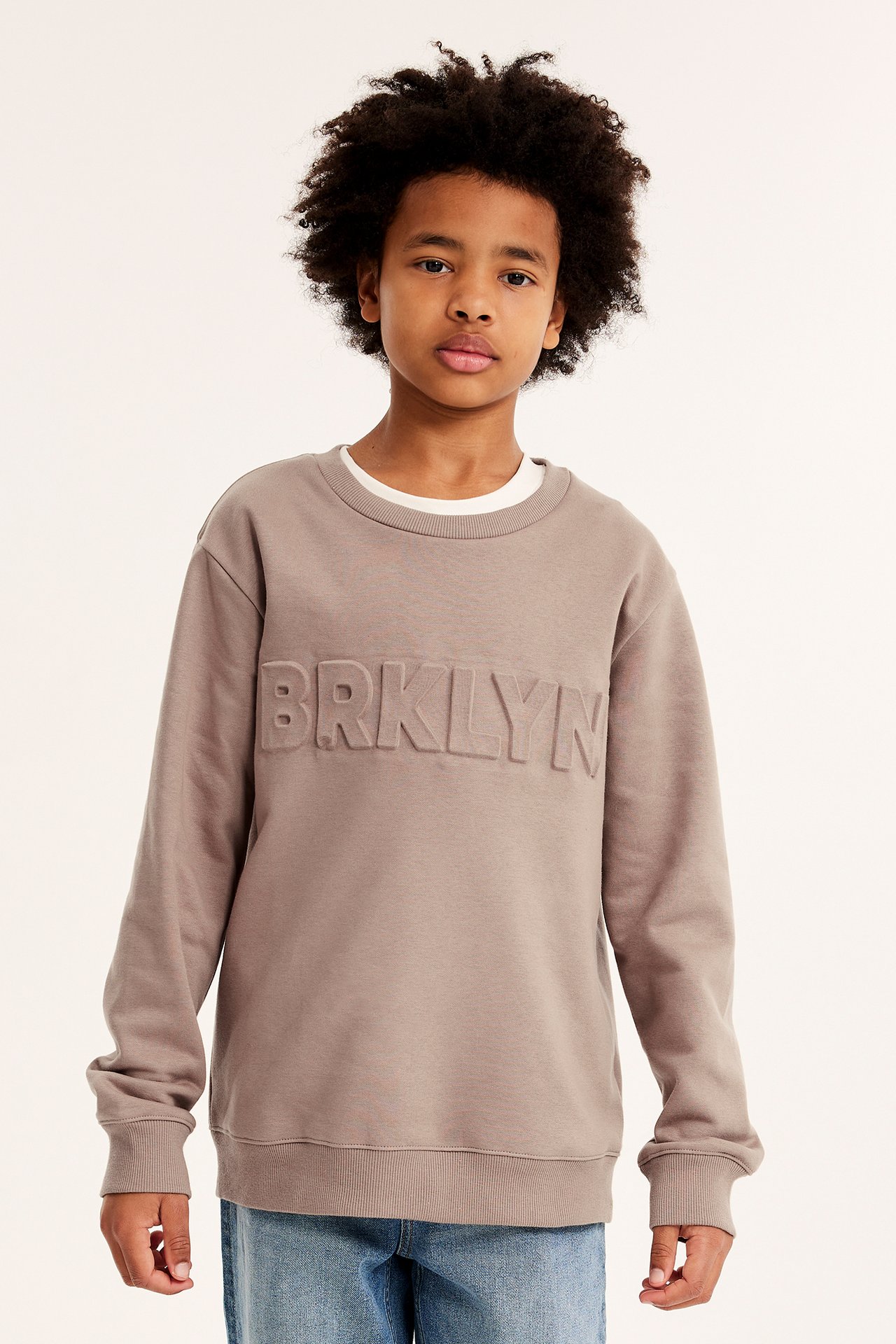 Frontvisning: barn i brun 'BRKLYN' sweatshirt med preget trykk over hvit t-skjorte og jeans.