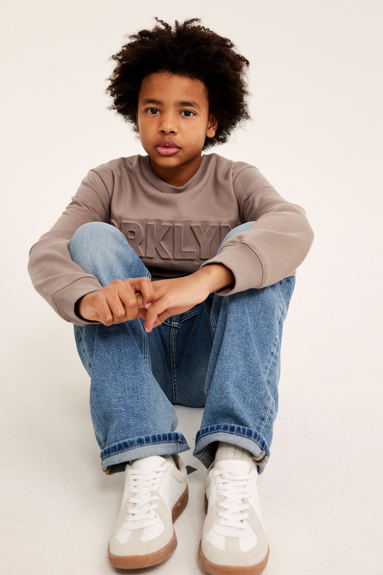 Frontbilde av et barn i brun sweatshirt med preget 'BRKLYN'-trykk, blå jeans og hvite joggesko.