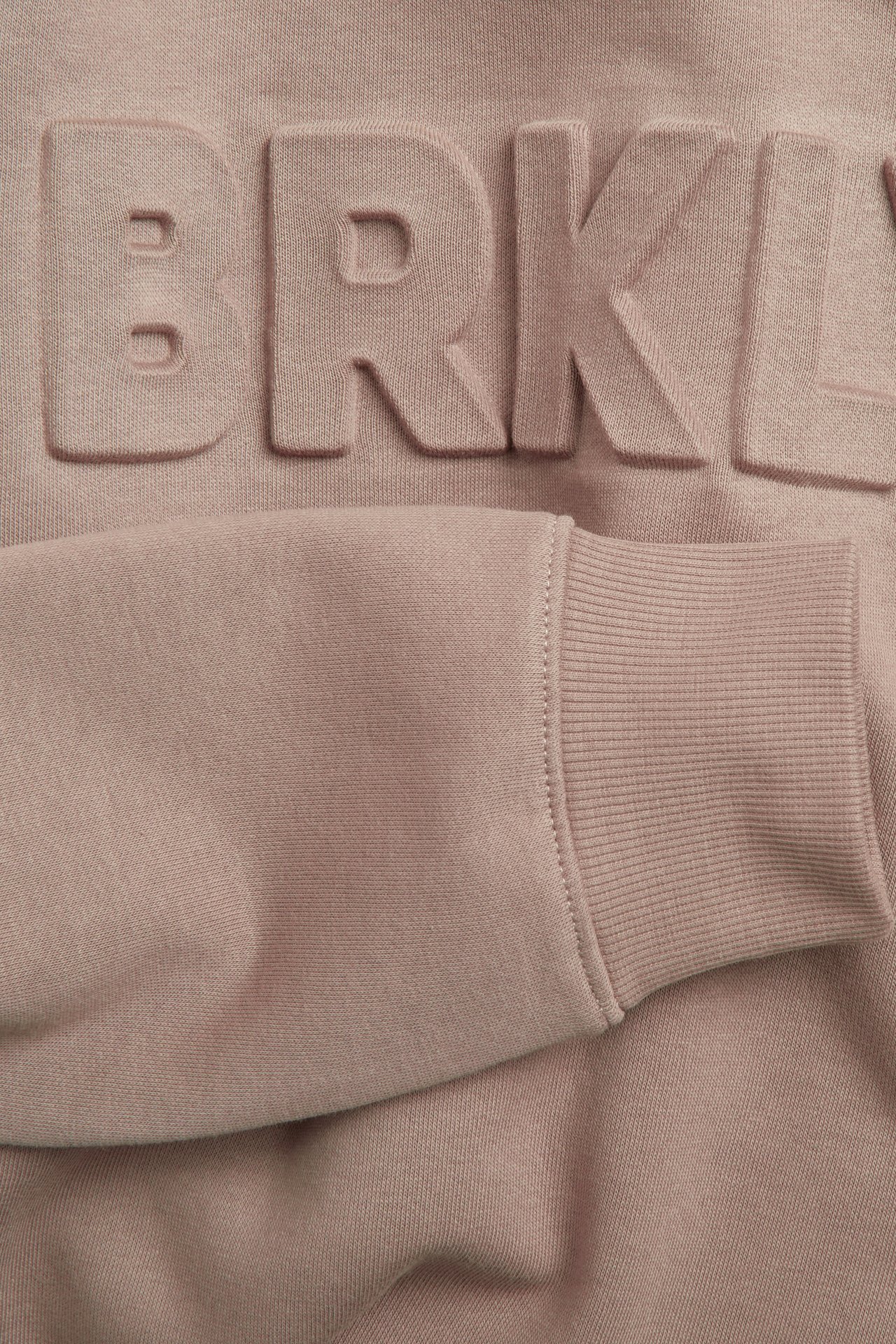 Nærbilde av en lys brun sweatshirt med preget 'BRKL'-trykk og ribbet mansjett.