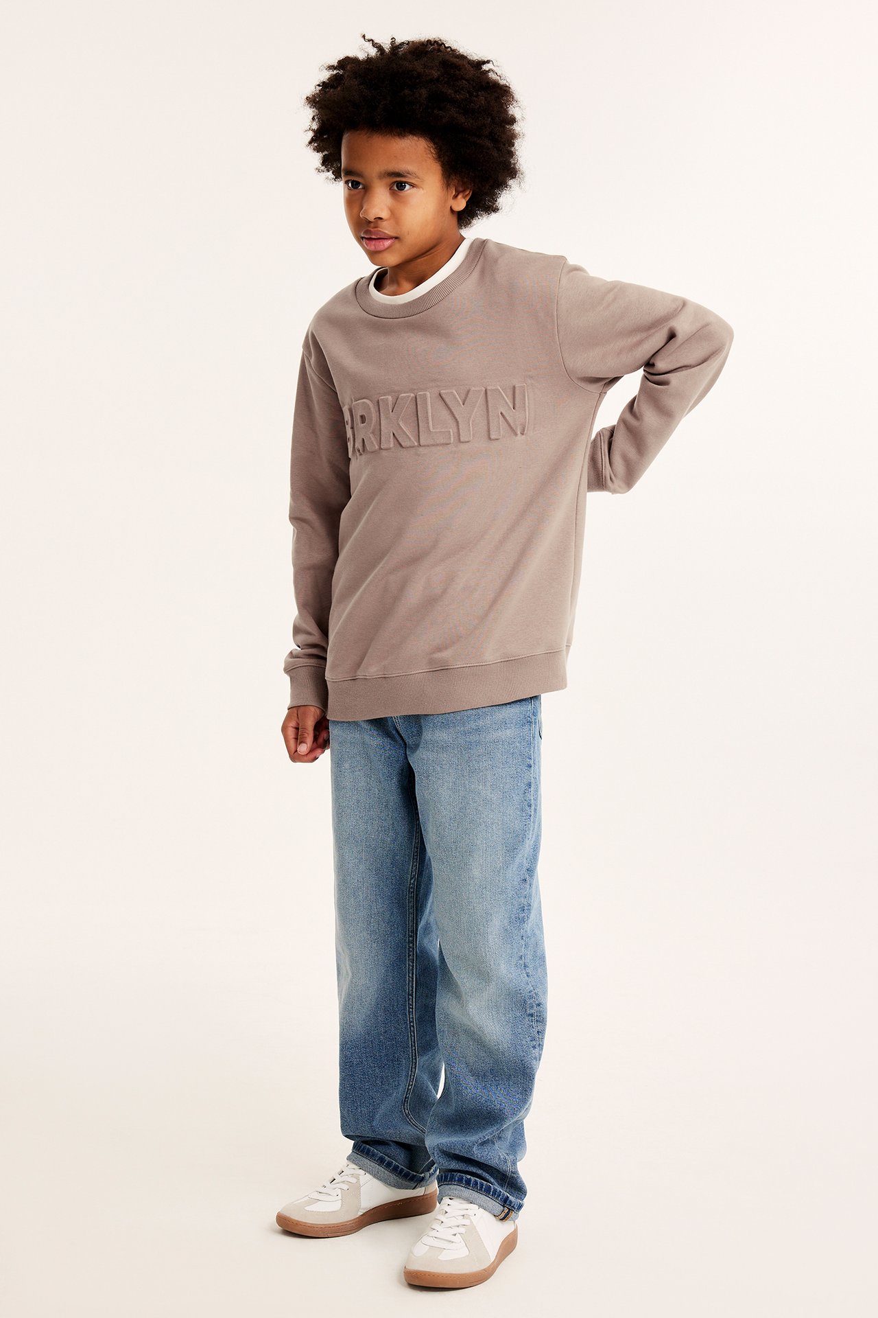 Frontbilde av et barn i brun sweatshirt med preget "BROOKLYN"-trykk, blå jeans og hvite joggesko.