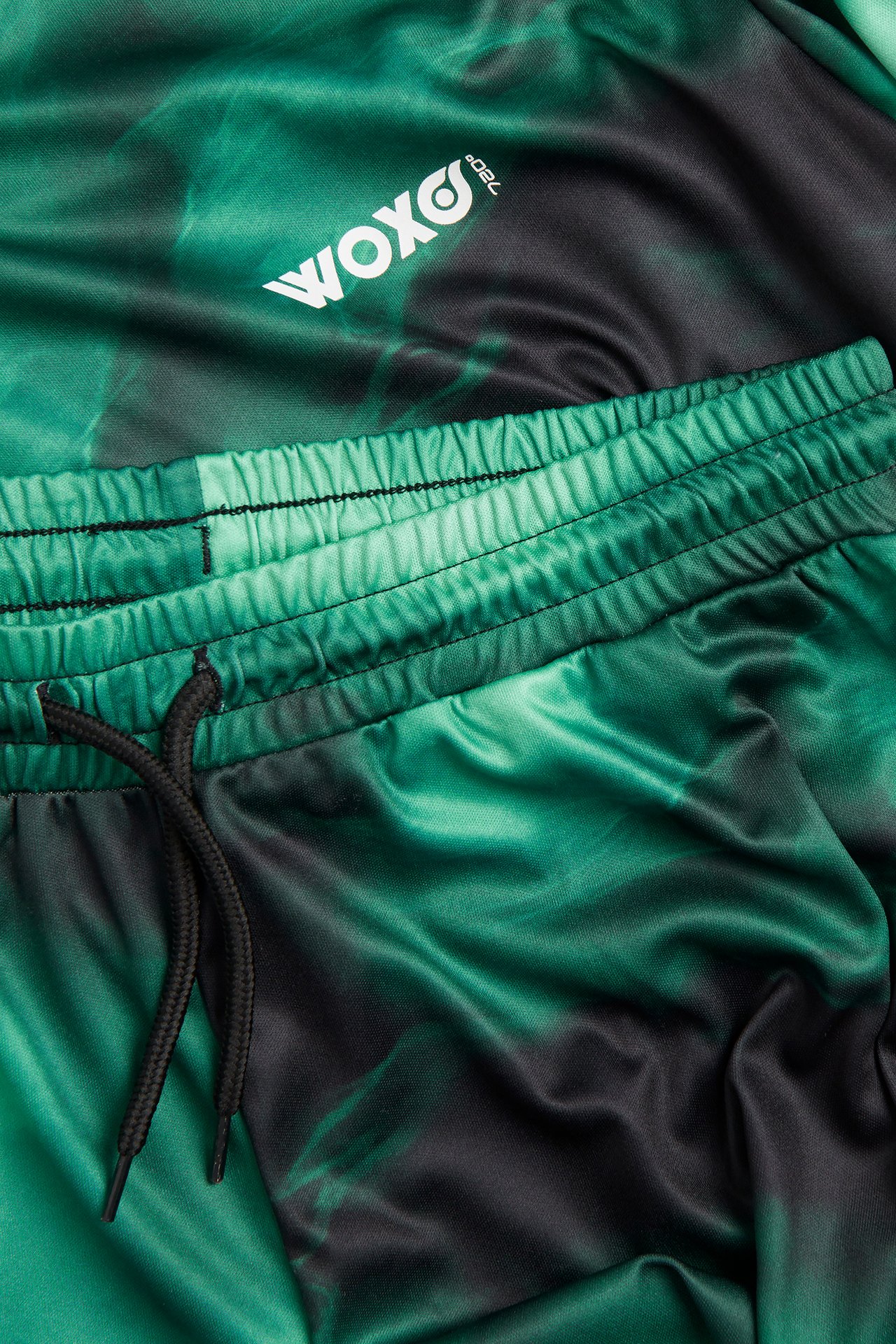 Närbild av ett mörkgrönt och svart mönstrat sportset i polyester med resår i midjan och vit logga.
