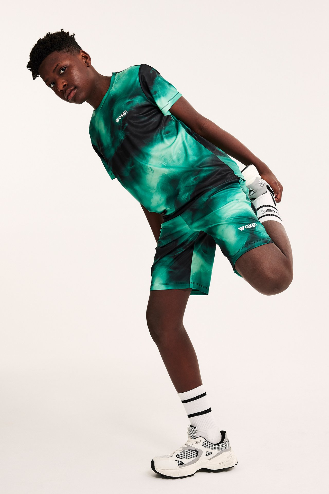Framifrån: Kid i mörkgrönt mönstrat sportset i polyester, vita strumpor, vita/grå sneakers, sträcker sig.
