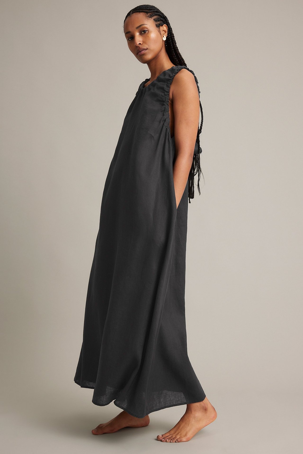 Sidovy av kvinna i lös svart ärmlös linneklänning med rynkad halsringning och sidfickor.