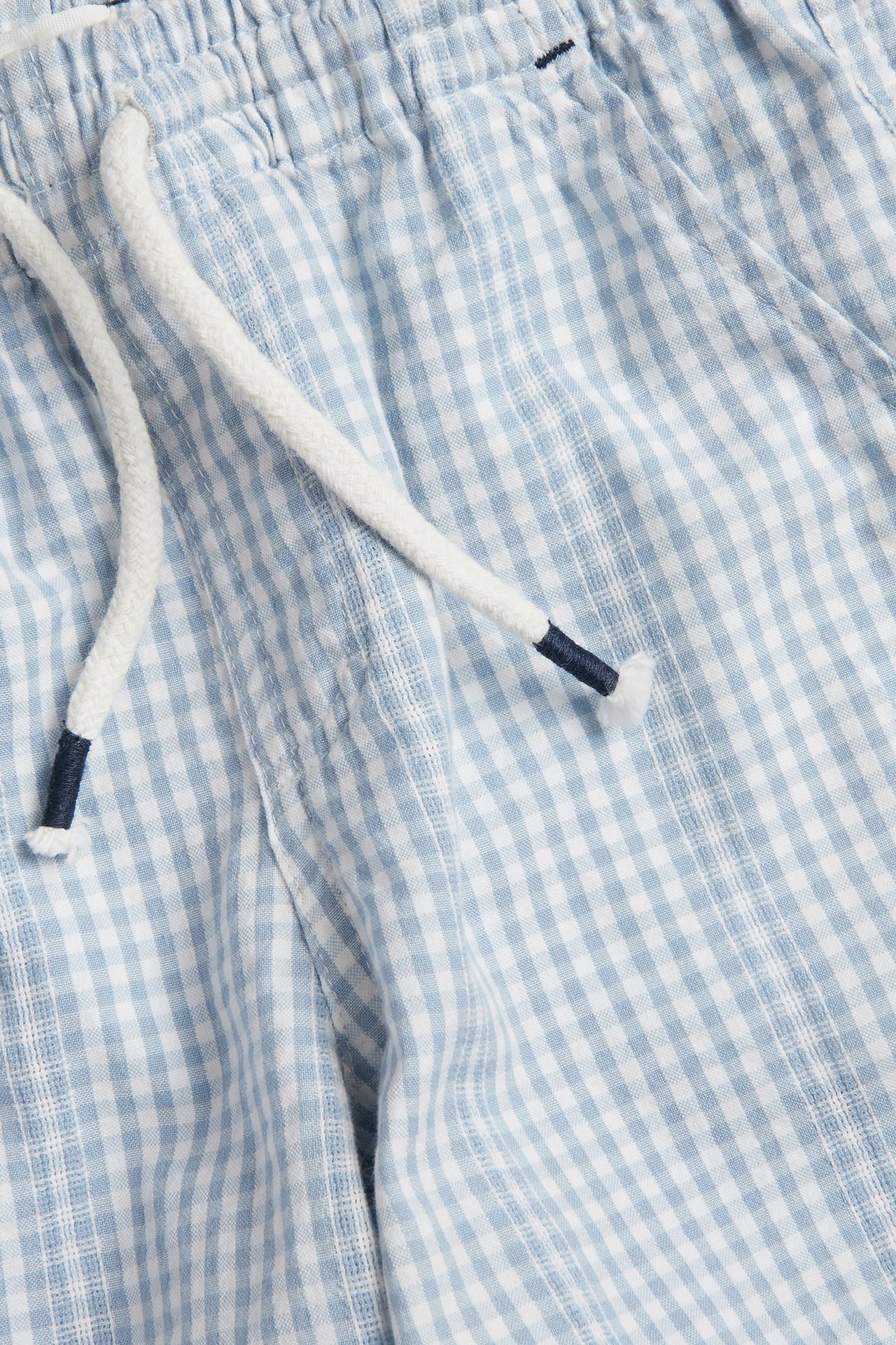 Nærbilde av lyseblå og offwhite rutete bomullsshorts med elastisk linning og hvit snøring.