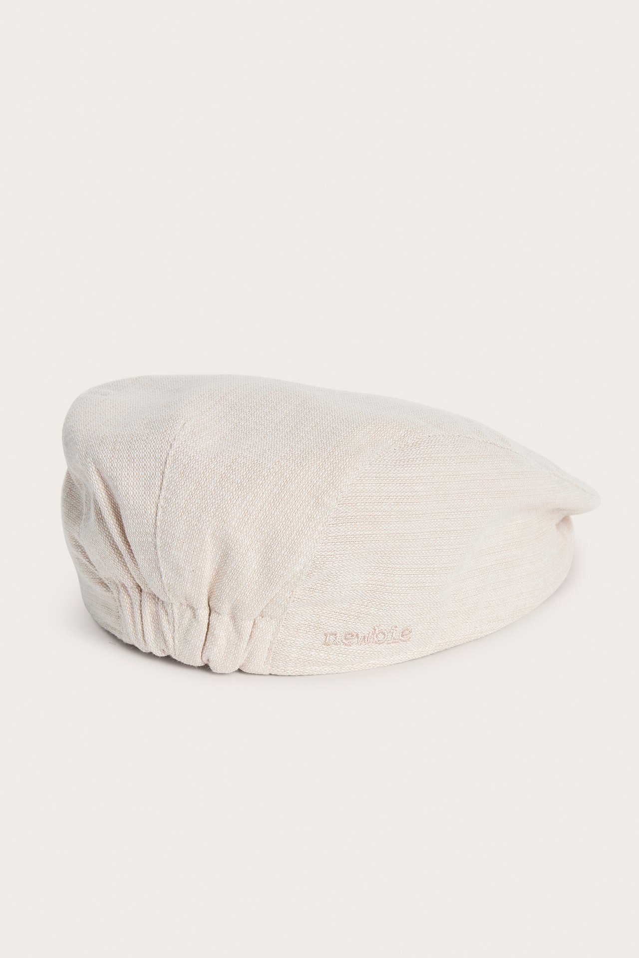 Sidovy av en beige texturvävd gubbkeps för baby/barn med resår bak och Newbie-broderi.