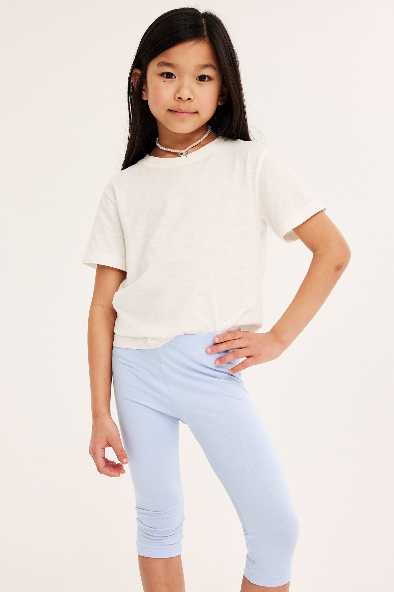 Widok z przodu: dziecko w jasnoniebieskich legginsach capri, białym T-shircie i naszyjniku z pereł.