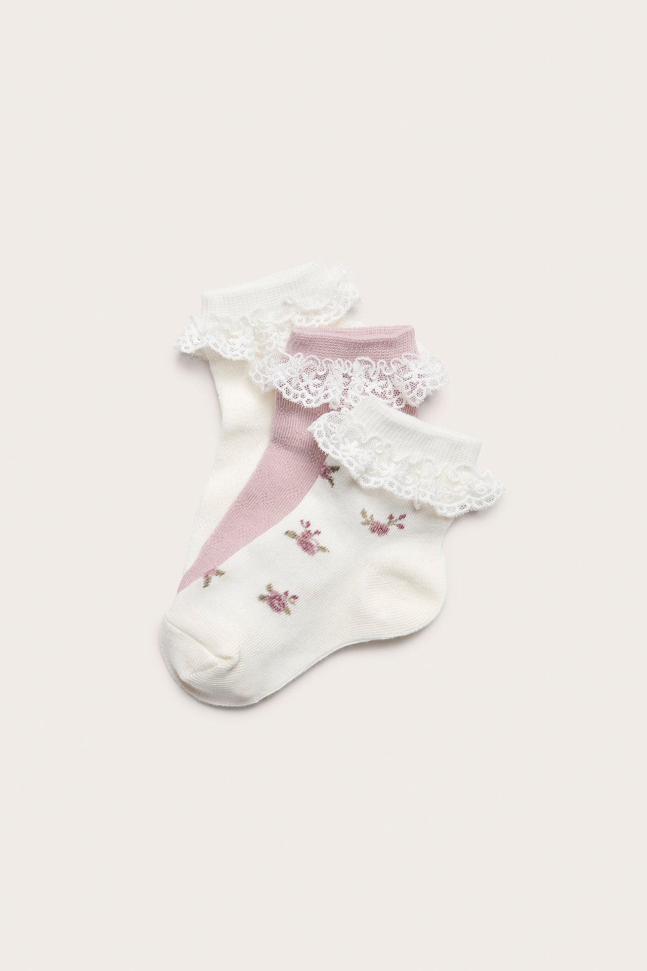 Framifrån: babysockor, en offwhite blommig, en offwhite pointelle, en ljusrosa pointelle, med vita volanger.