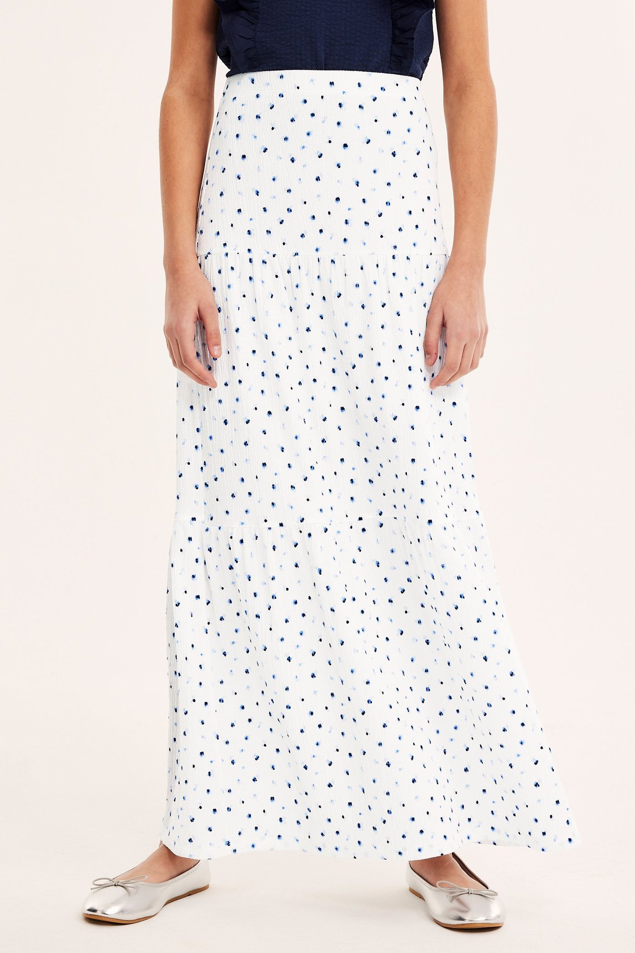 Frontvisning av et barn i et hvitt blomstret krøllet jersey maxiskjørt, en mørkeblå topp og sølvfargede ballerinasko.