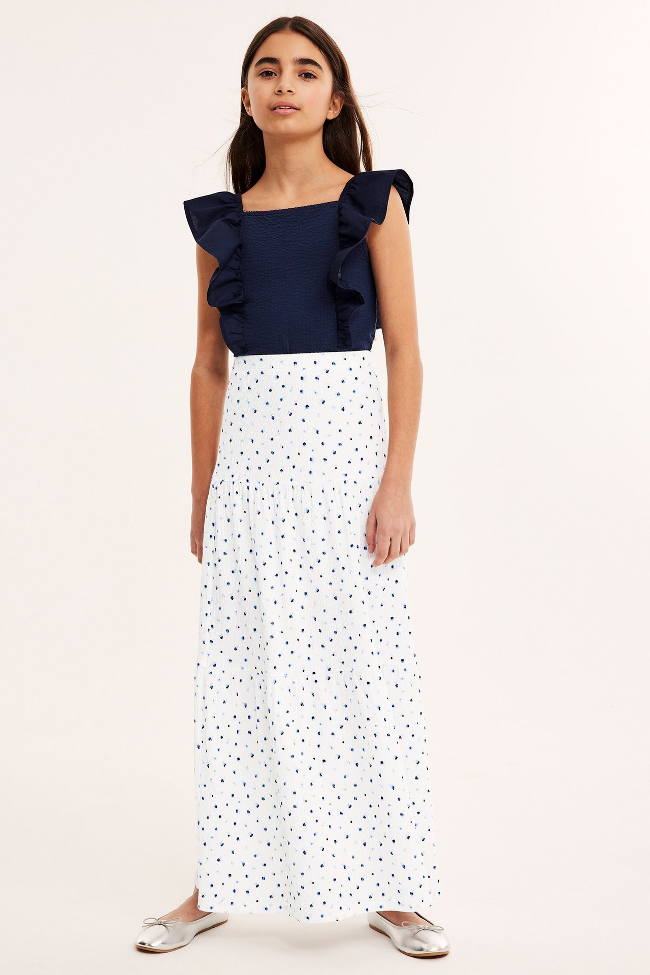 Frontalvisning: Barn i hvitt mønstret maxiskjørt, marineblå volangtopp og sølvfargede ballerinasko.