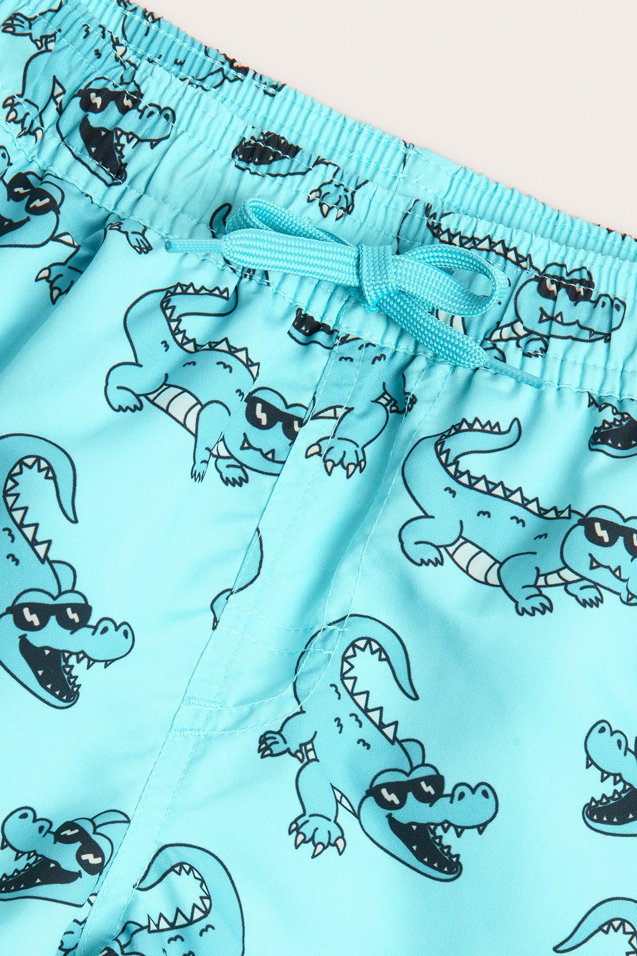 Närbild på turkosa badshorts för barn med krokodil- och solglasögonmönster, resår i midjan, dragsko.