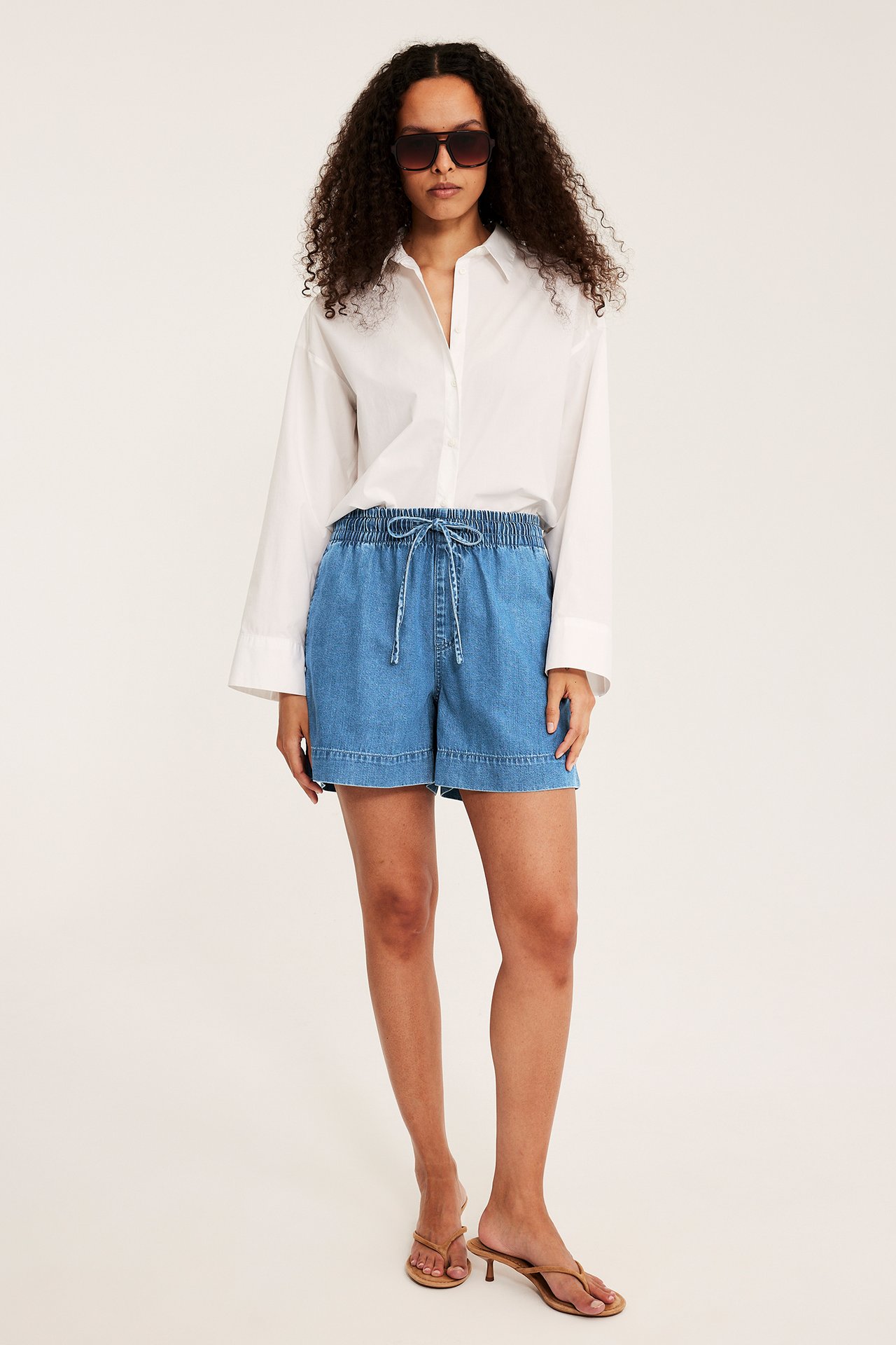 Widok z przodu kobiety w niebieskich jeansowych szortach pull-on ze sznurkiem, białej koszuli i okularach.