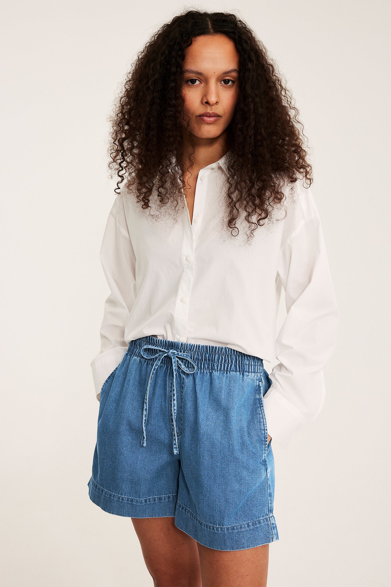 Widok z przodu: kobieta w niebieskich szortach jeansowych pull-on z troczkiem i białej koszuli.