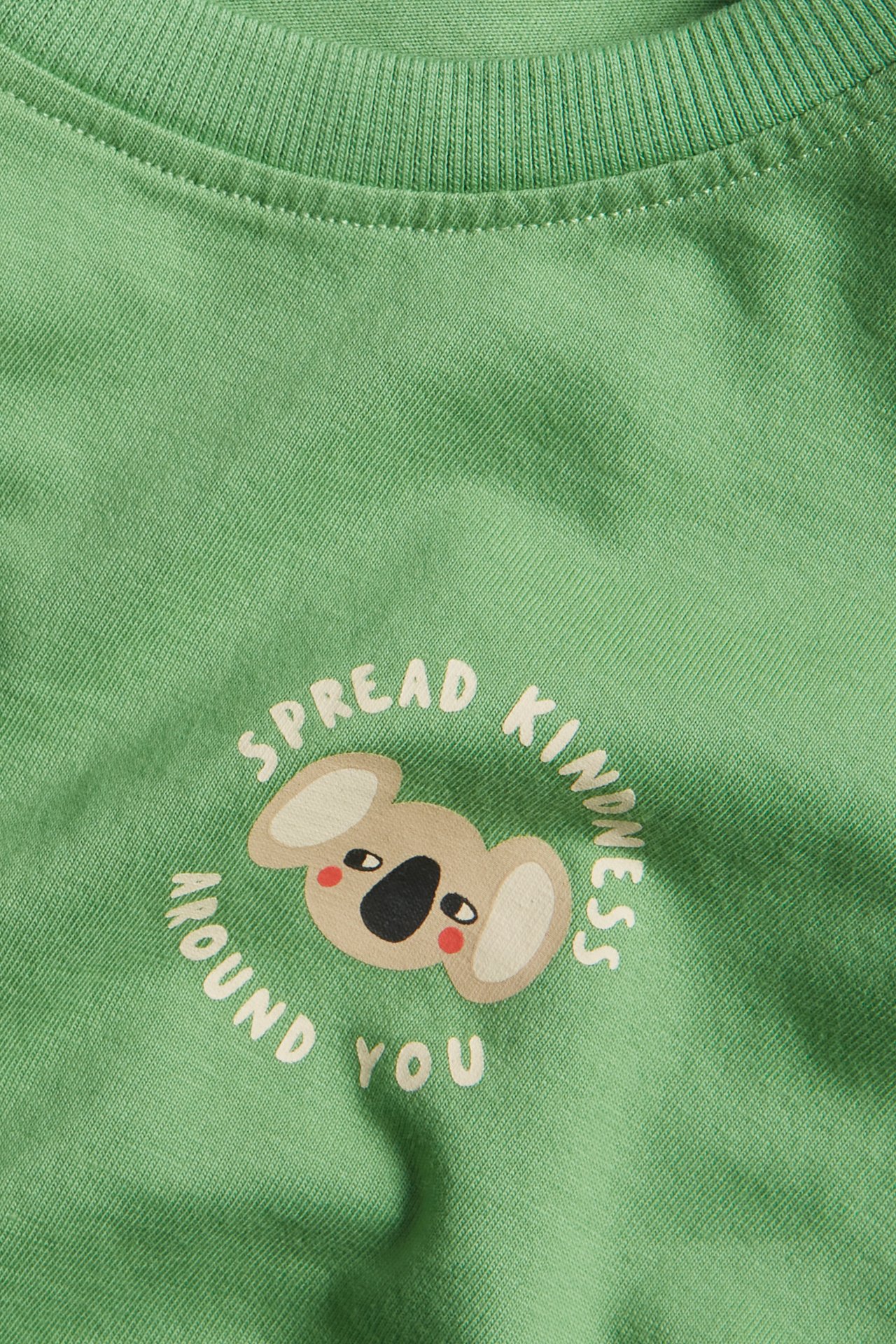 Lähikuva lasten vihreästä puuvilla-T-paidasta, jossa koala- ja "Spread Kindness" -painatus.