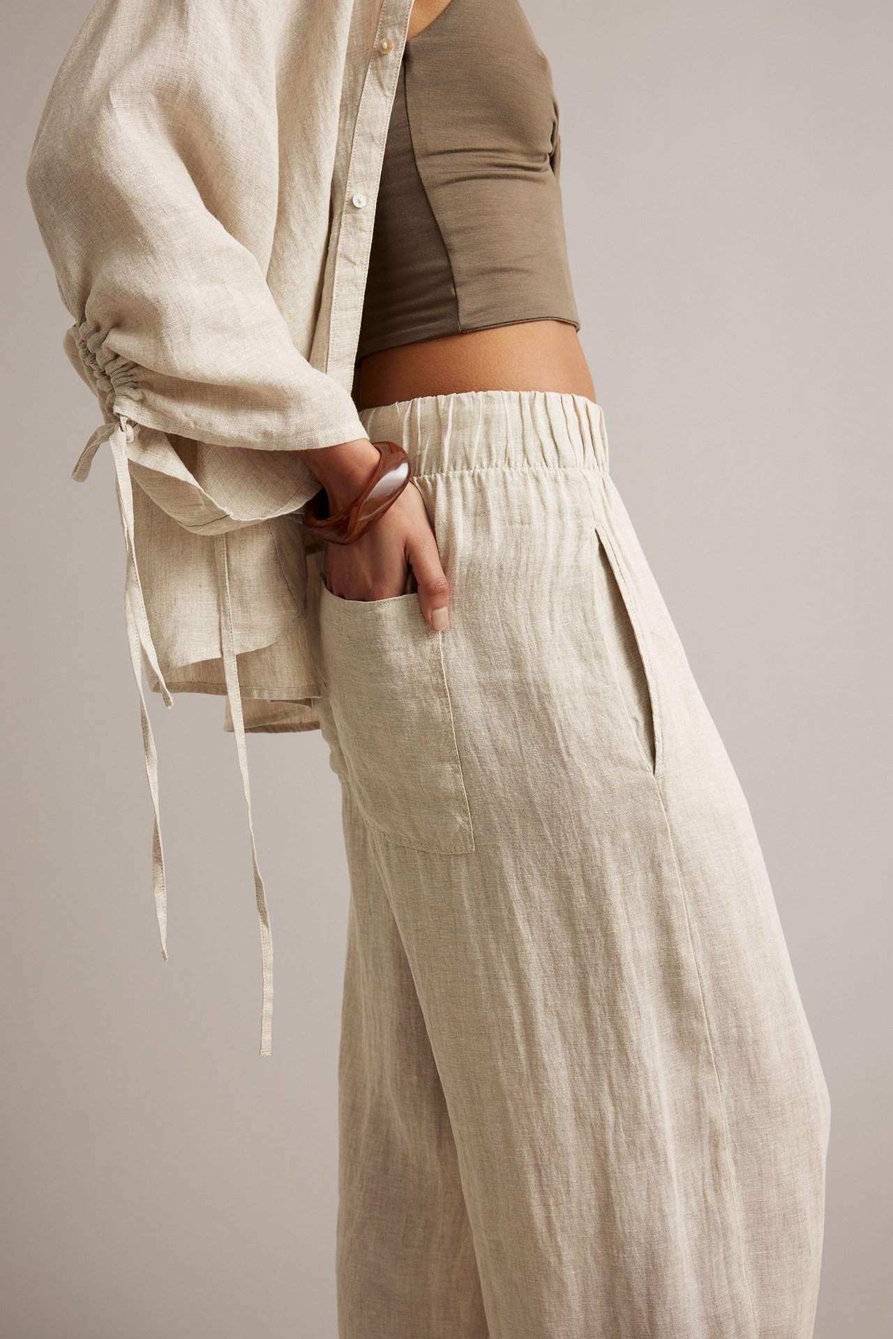 Sidovy: Kvinna i beiga melerade linnebyxor med barrel-fit och resår i midjan. Bär brun croppad topp och beig linneskjorta.