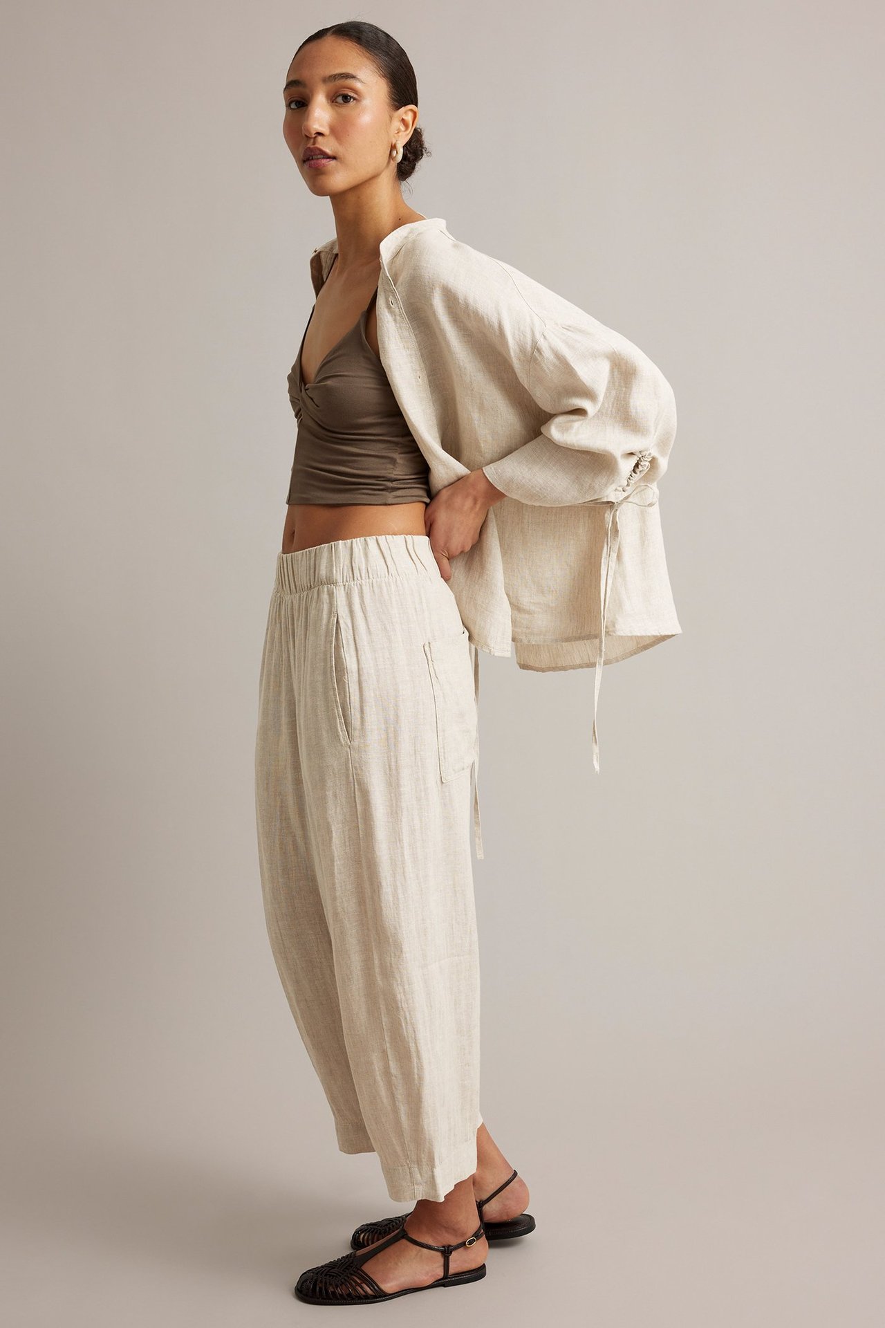 Sidovy: kvinna i beige melerade linnebyxor, olivfärgad croptop och ljusbeige linneskjorta.