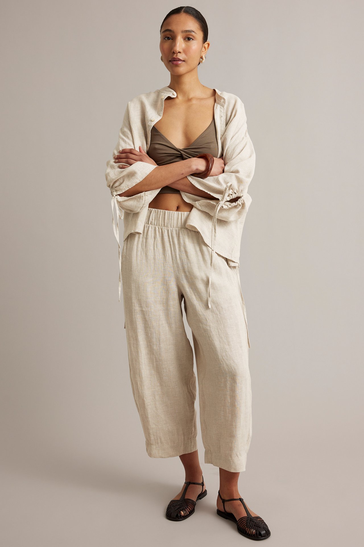 Framifrån: Kvinna i beigemelered barrel-leg linnebyxor, öppen beige linneskjorta och olivfärgad topp.