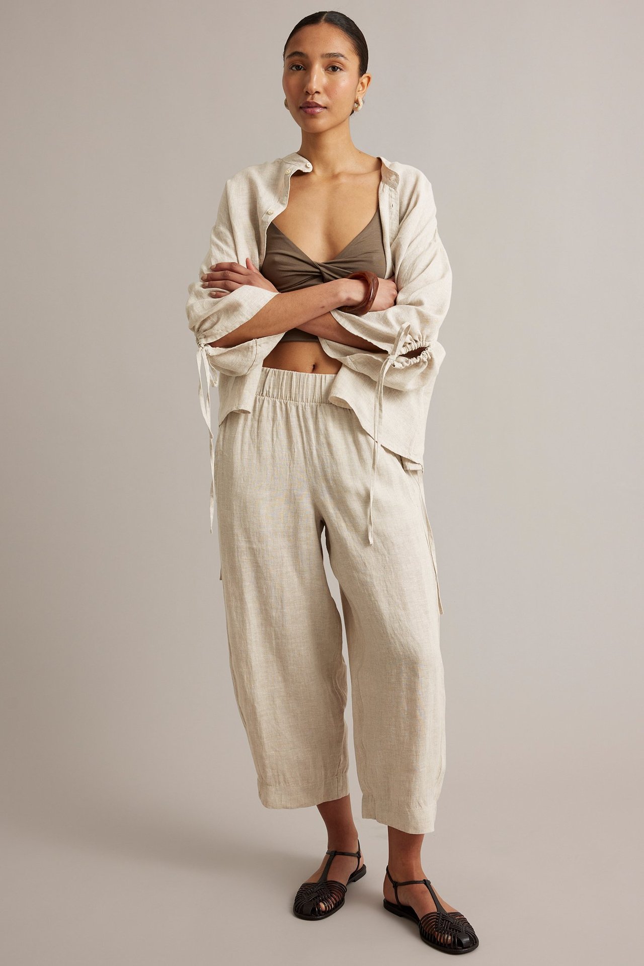 Edestä: Nainen yllään beigenmelatut barrel-leg pellavahousut, avoin beige pellavapaita ja oliivinvärinen toppi.