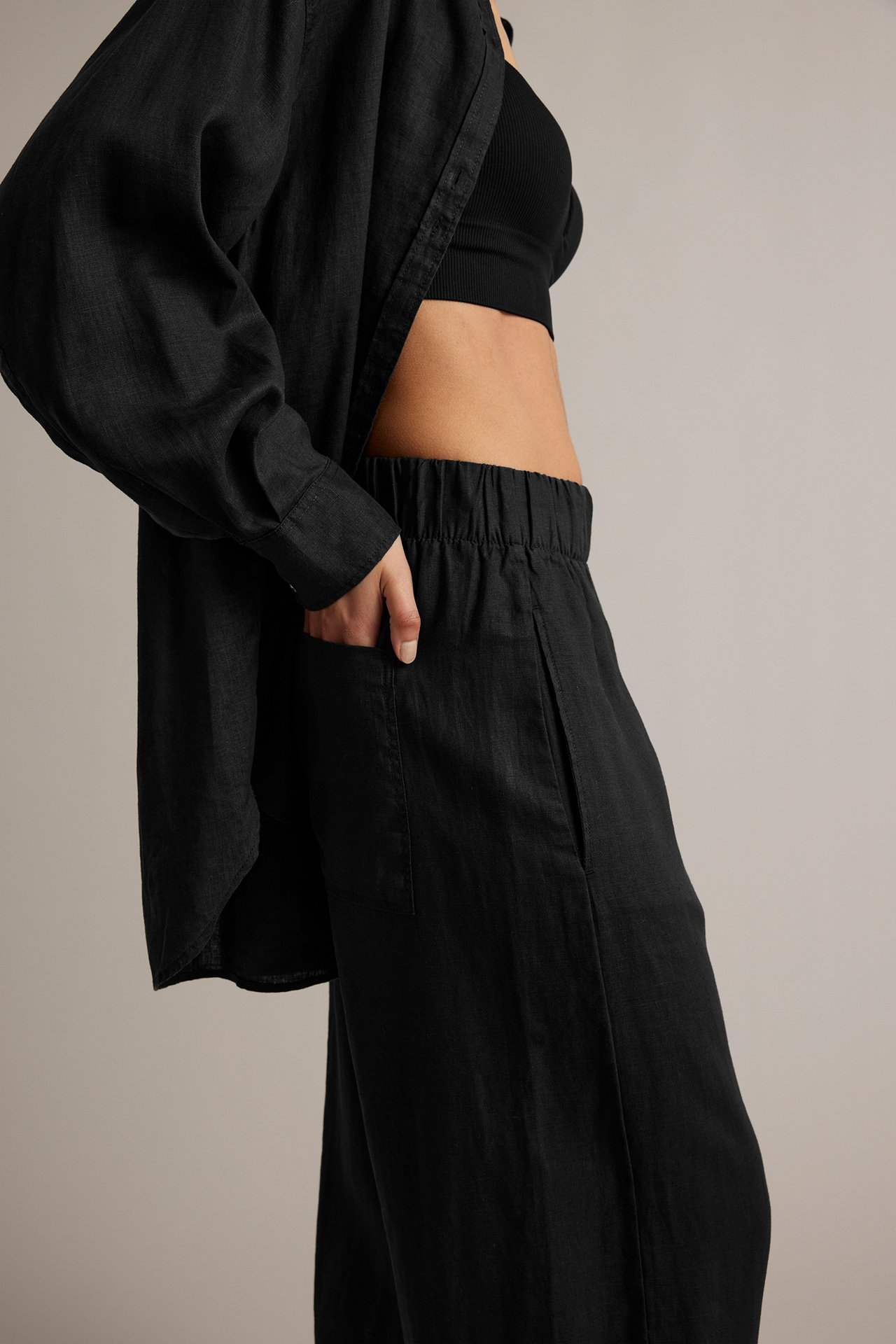 Sidebilde av kvinne i svart linbukse med barrel leg, elastisk midje og hånd i lommen, med svart ribbet topp og åpen linskjorte.