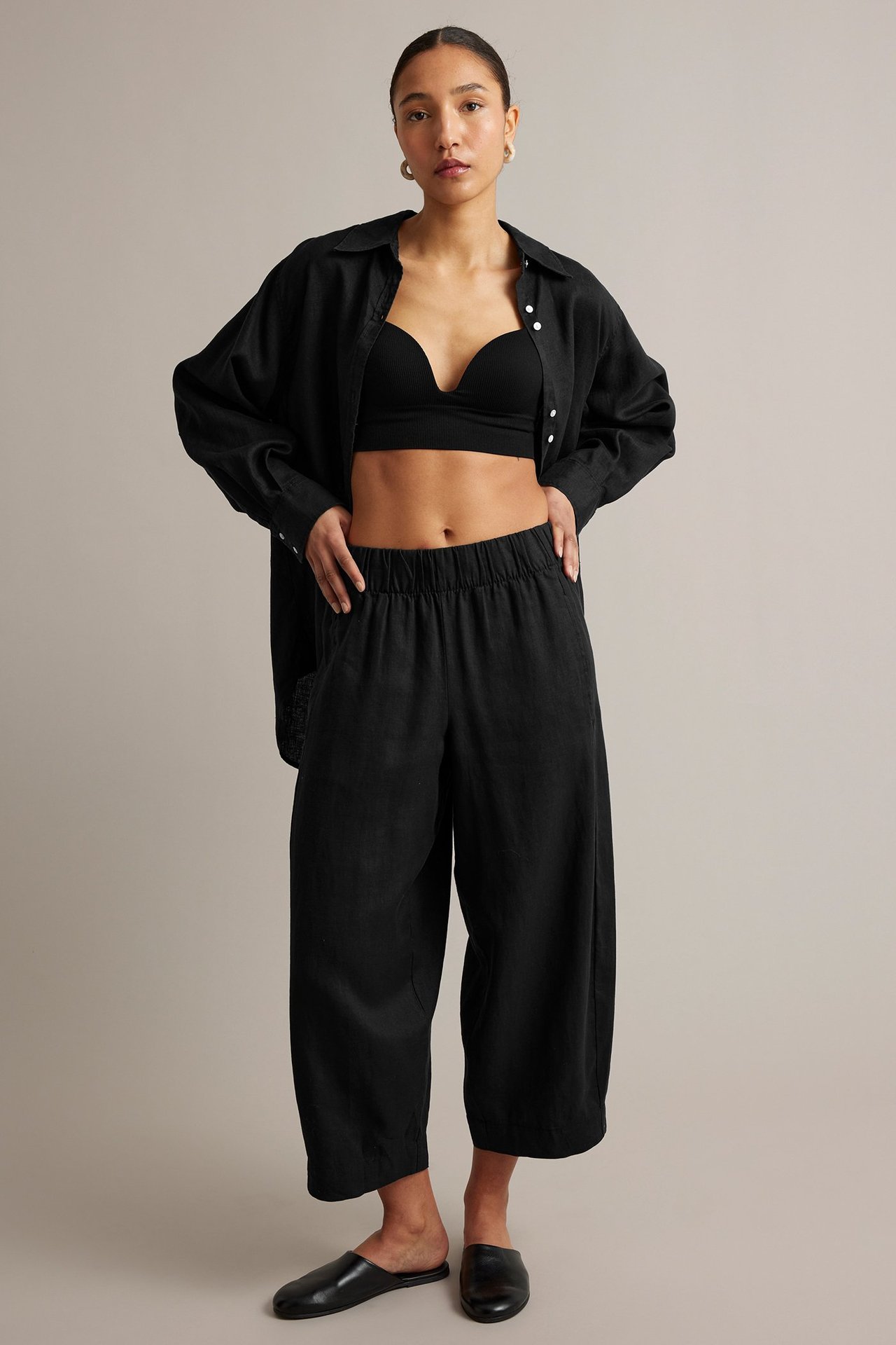 Frontbilde av kvinne i svarte barrel fit linbukser, svart BH-topp, åpen linskjorte og svarte slip-on sko.