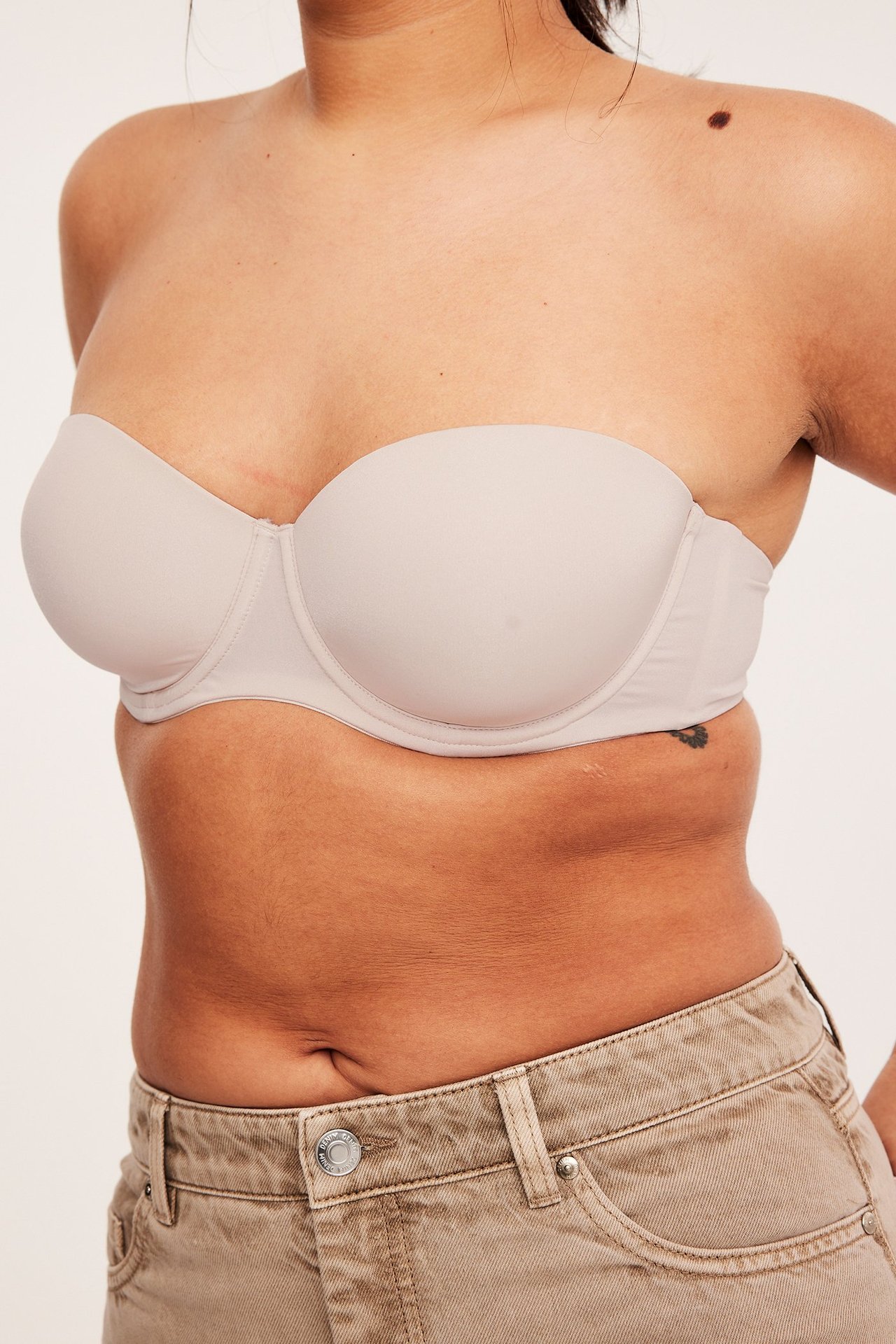 Närbild framifrån på en kvinna i en beige axelbandslös balconette-bh och beige jeans.