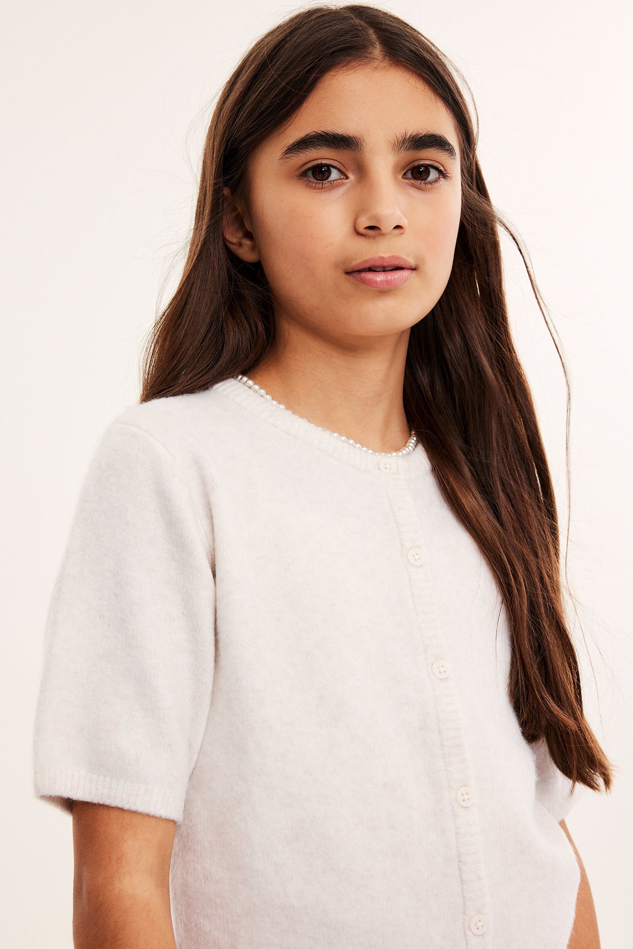 Lähikuva edestä lapsesta, jolla on valkoinen lyhythihainen neuletakki helmikoristeella ja nappikiinnityksellä.