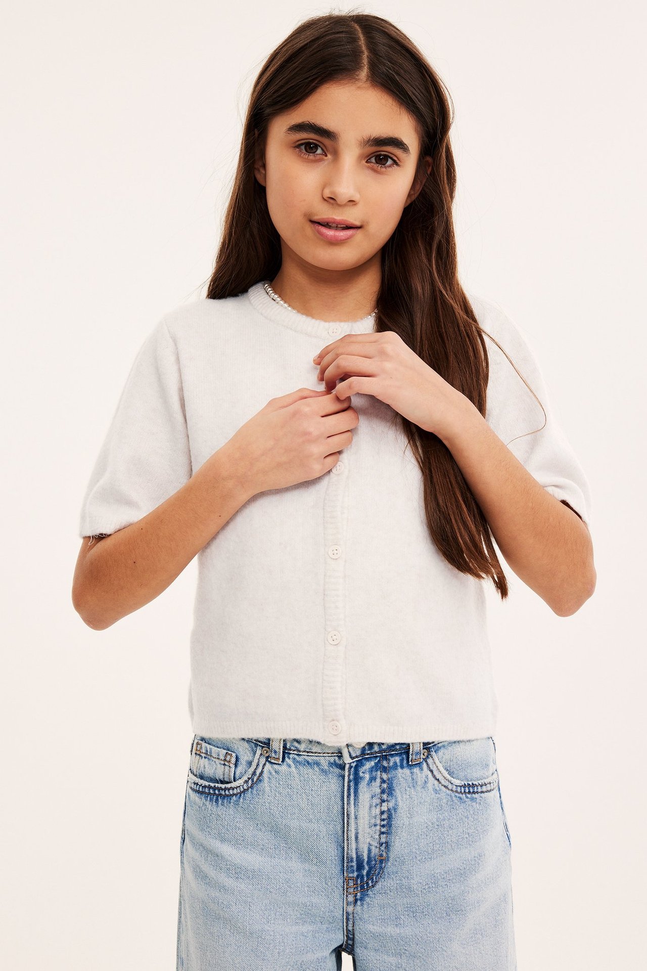 Edestä näkymä lapsesta, jolla on valkoinen lyhythihainen neuletakki napeilla, vaaleansiniset farkut ja helmikaulakoru.