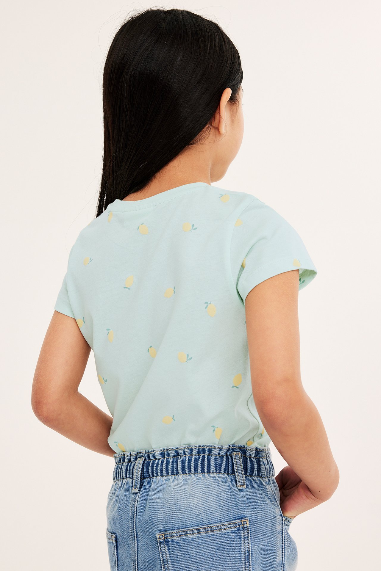 Bakifrån: ett barn i en ljusgrön kortärmad T-shirt med gult citronmönster och blåa jeans.