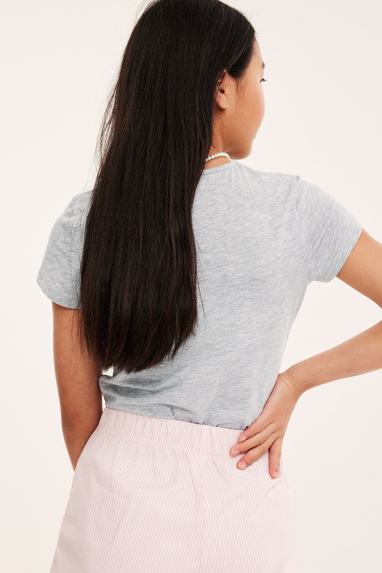 Bakifrån: ett barn i ljusgrå melerad T-shirt, rosa randiga shorts och ett pärlhalsband.
