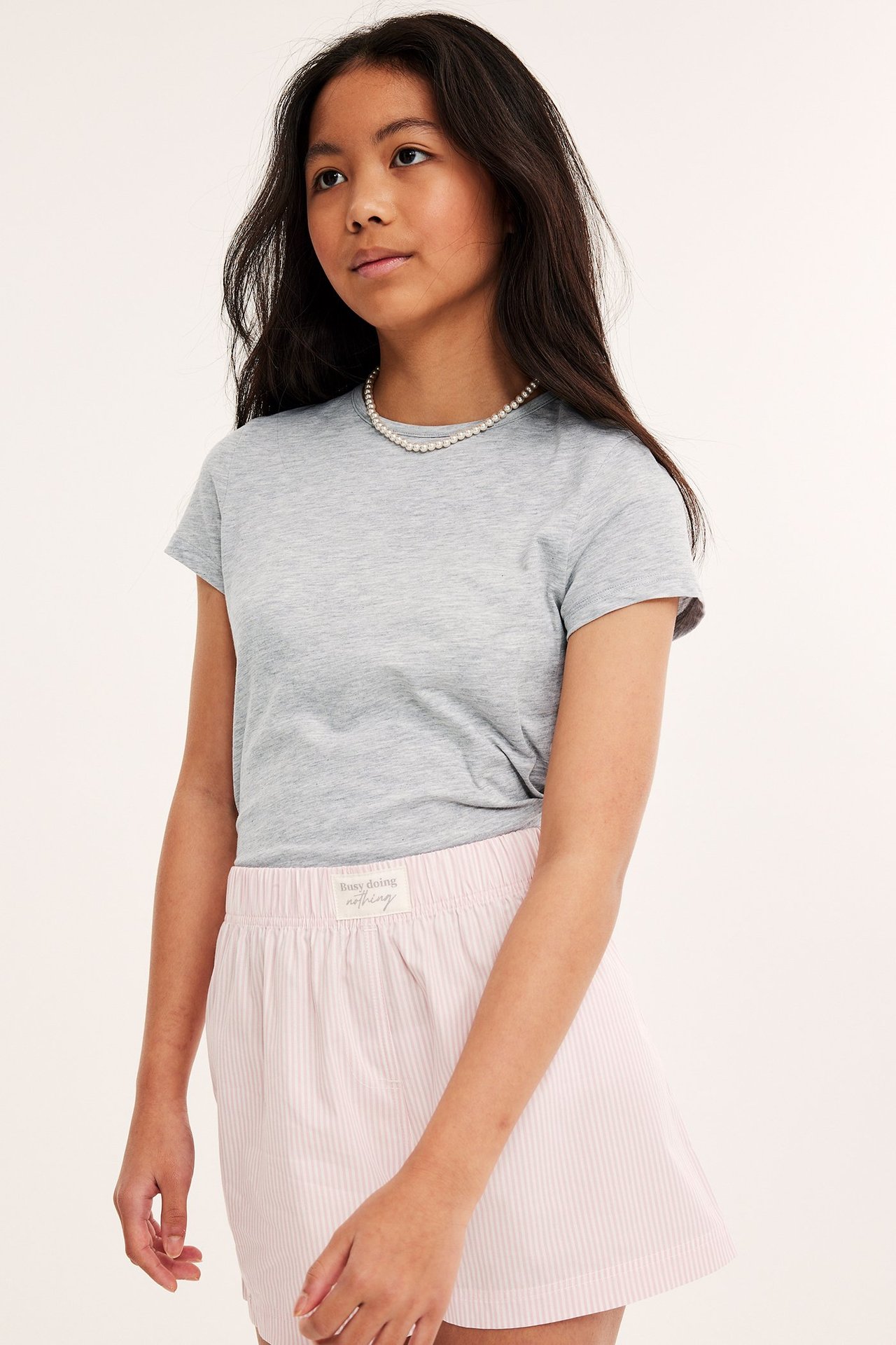 Framifrån: ett barn i ljusgrå melerad t-shirt, rosa randiga shorts och ett pärlhalsband.