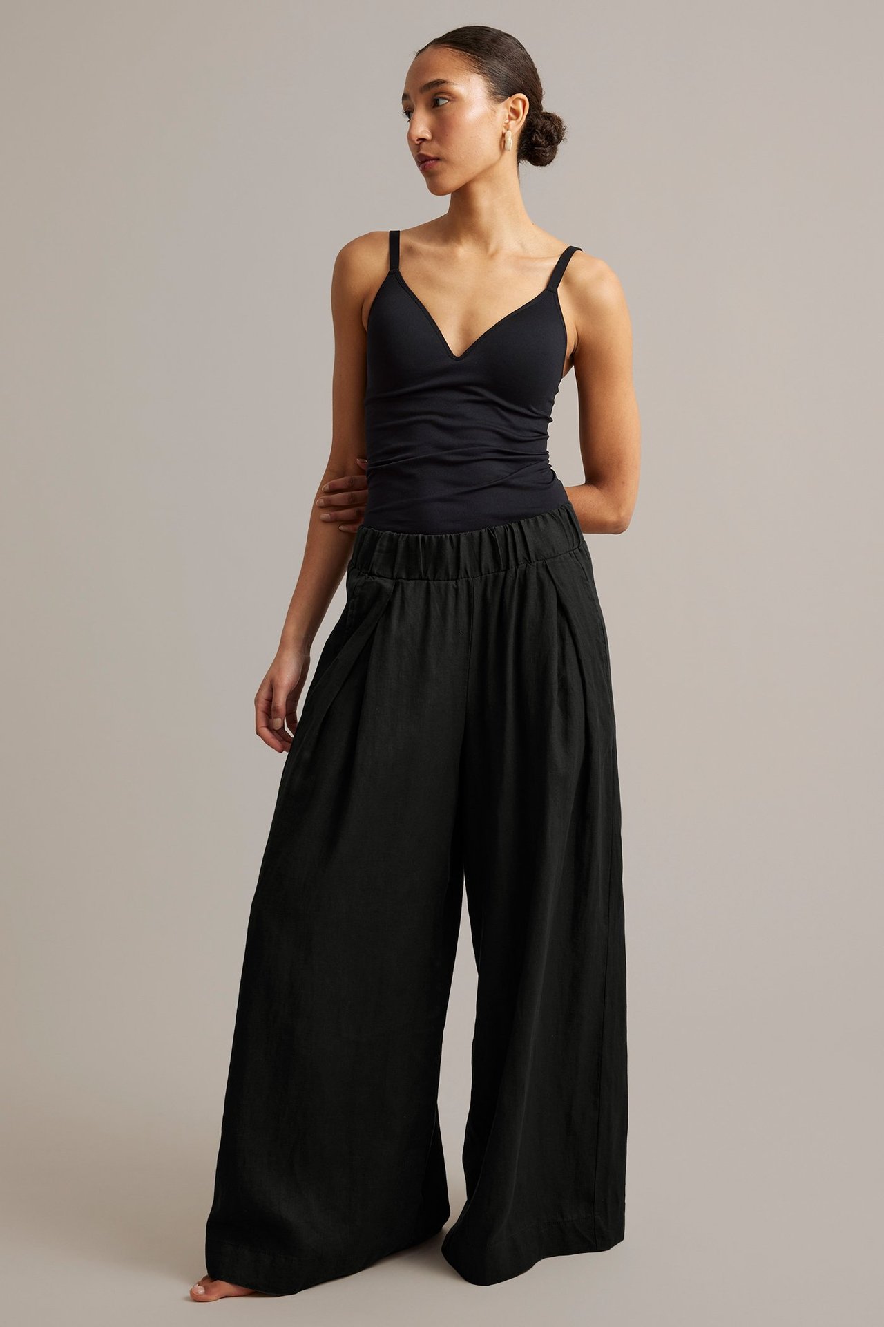 Naisen etunäkymä: mustat leveälahkeiset pellavahousut laskoksilla ja musta V-kauluksinen toppi.