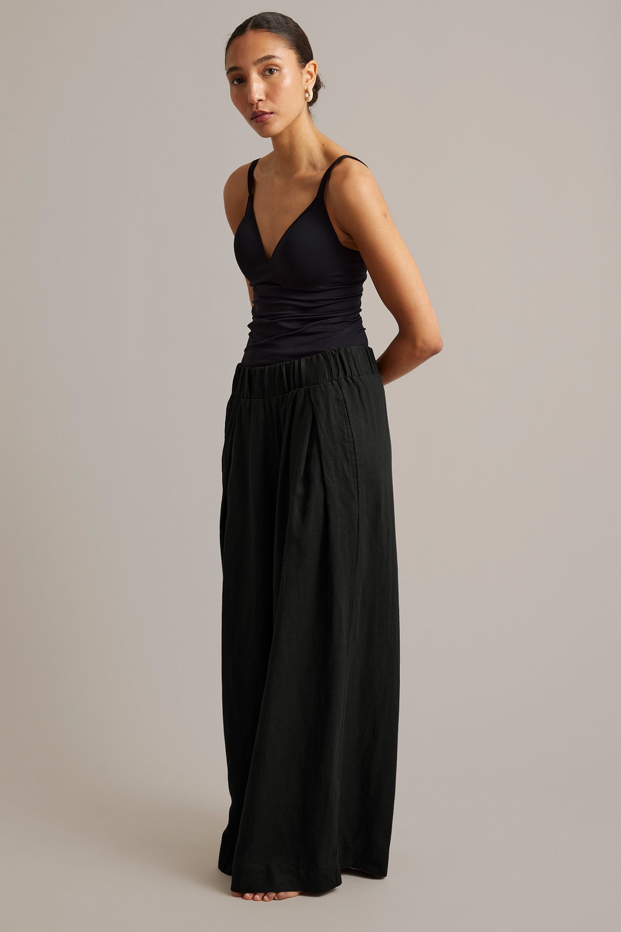 Edestä: Nainen mustissa väljälahkeisissa pellavahousuissa joustovyötäröllä ja laskoksilla, musta toppi.